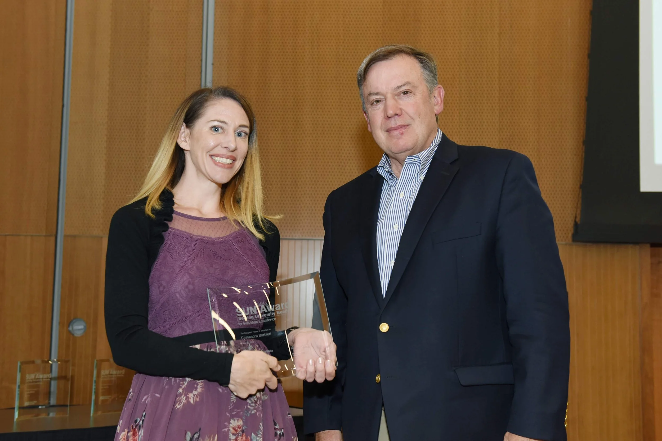 A woman and a man standing indoors, smiling, with the woman holding an award plaque. The woman has long, light brown hair and is wearing a purple dress with black sleeves. The man has short, gray hair and is dressed in a dark blazer with a striped shirt.
