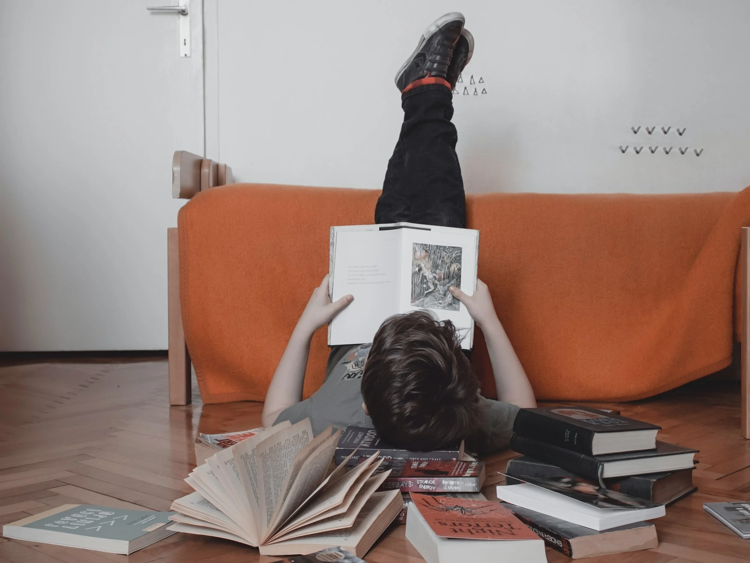 A child lying on the floor under a table, reading a book with feet propped up on the underside of the table, surrounded by scattered books and papers.