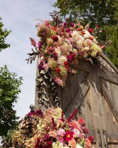 Large floral arrangement with pink, red, and white flowers on a rustic wooden structure outdoors.