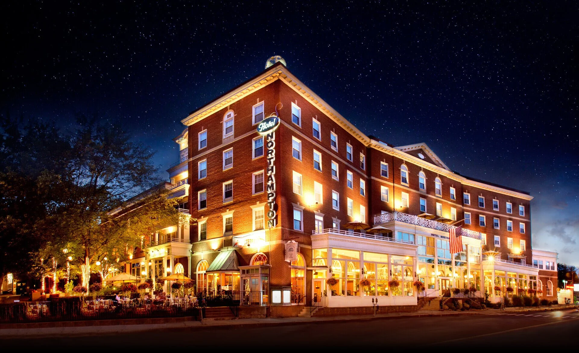 Nighttime view of the Hotel Northampton in Massachusetts with bright exterior lights. The sky above is filled with stars, and the hotel's architecture features brick and white accents with outdoor seating and flags in front.