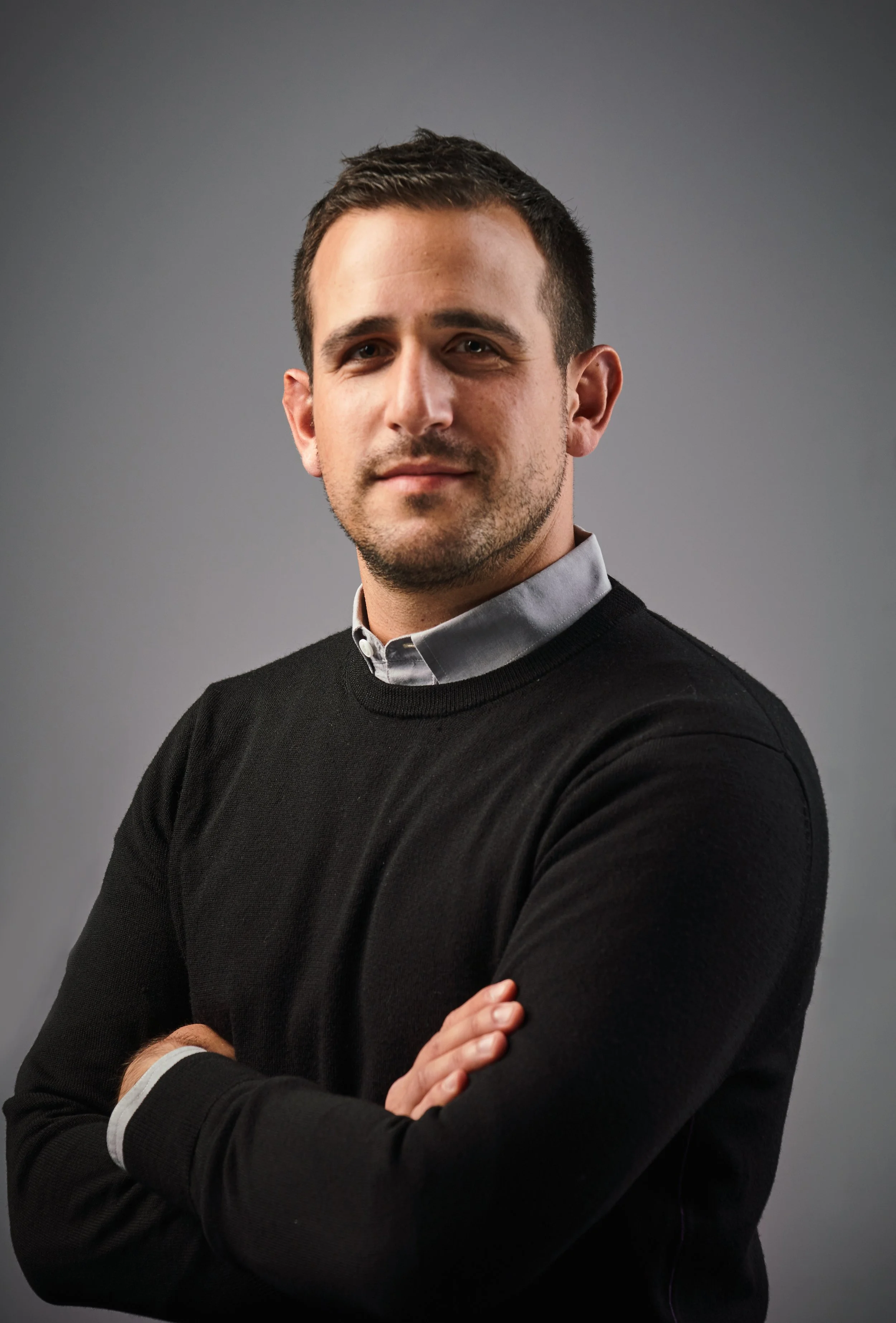 Portrait of founder, Steven Lemeshow, wearing a black sweater over a collared shirt, crossing his arms, standing against a plain gray background.