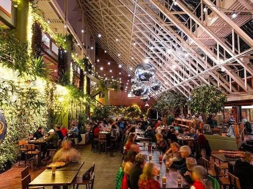 Indoor restaurant with high ceiling, string lights, large hanging globe sculptures, greenery, and many diners at tables.
