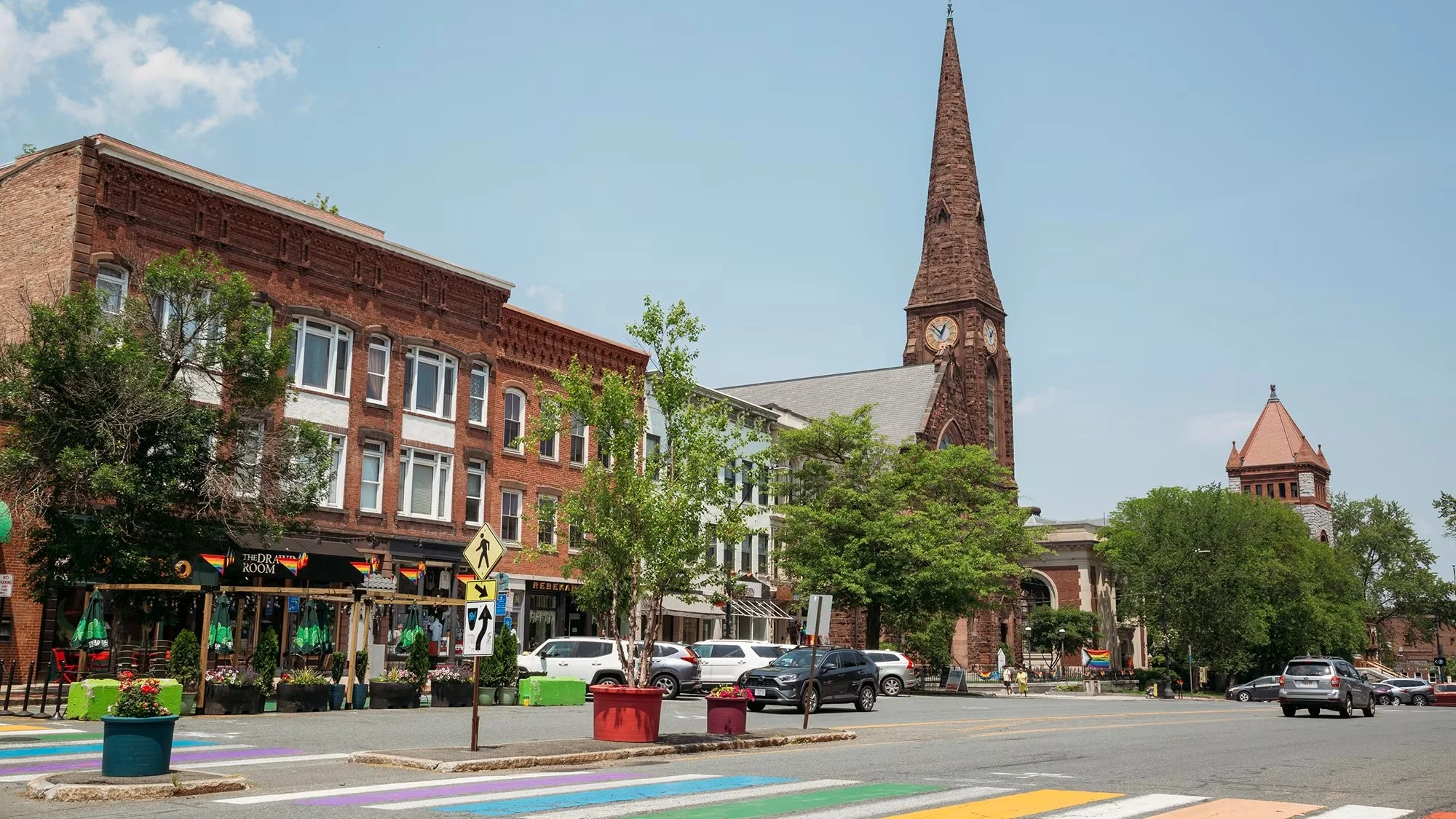 City street scene with historic brick buildings, a church with a tall steeple, trees, parked cars, outdoor seating, rainbow crosswalk, and pedestrian signs.
