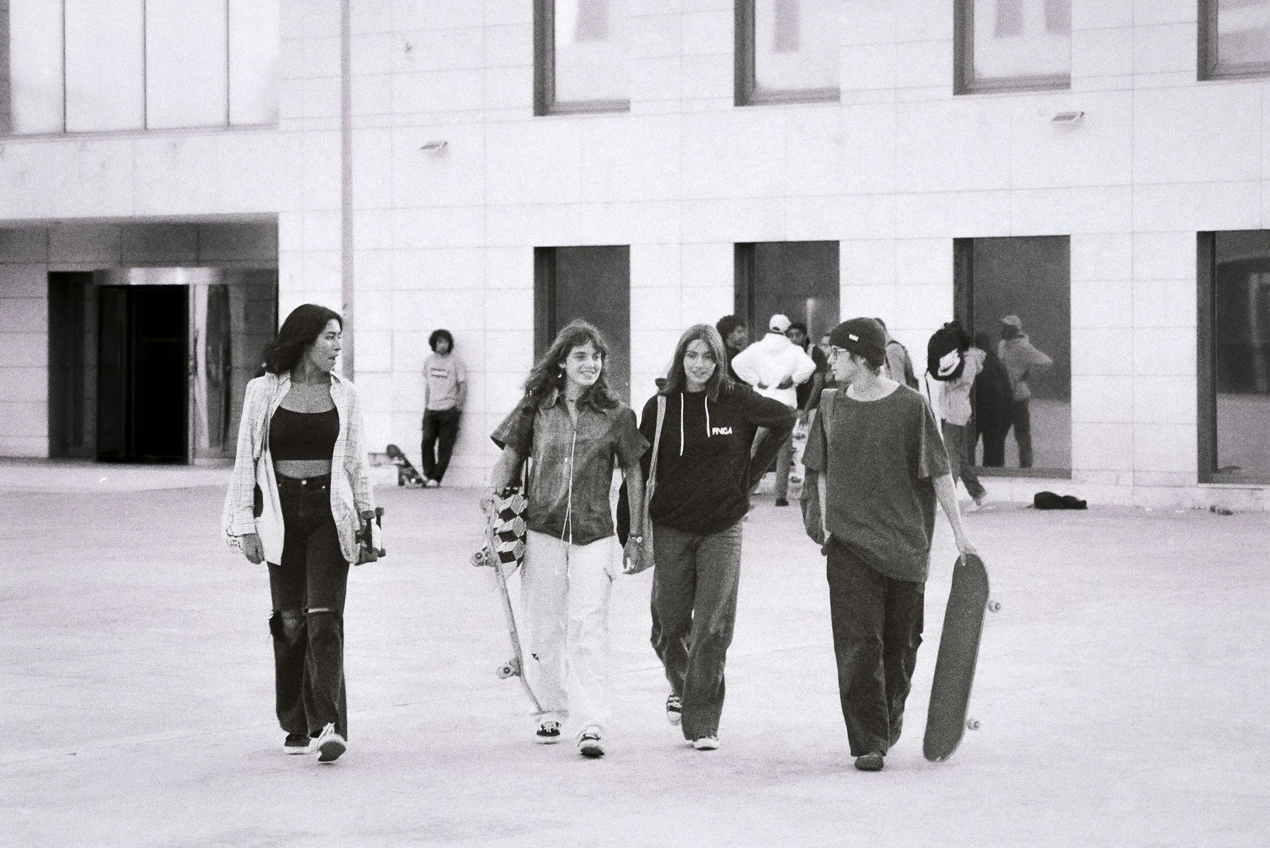 Black and white photo of four young adults walking confidently together, carrying skateboards, in an open urban plaza