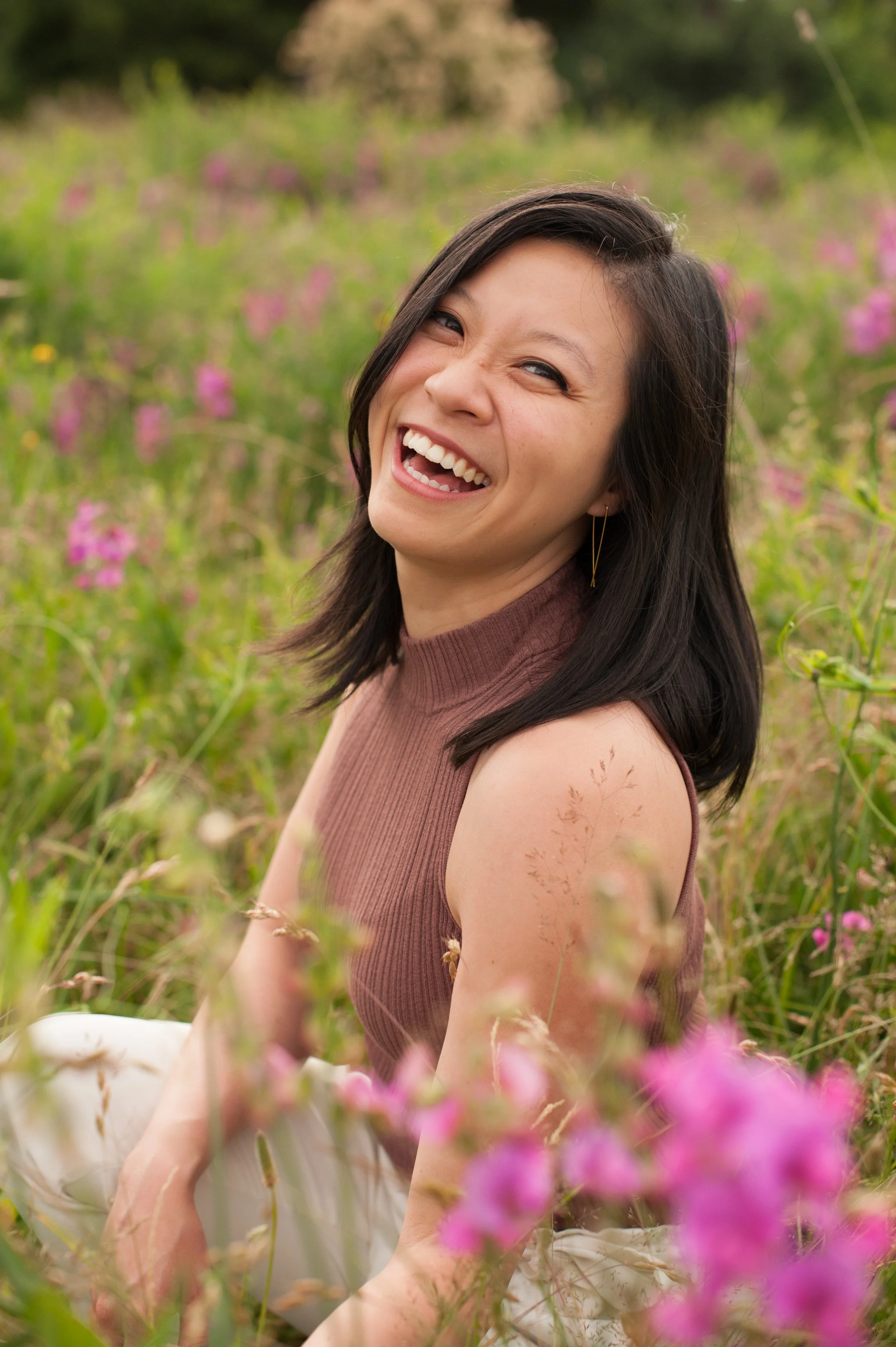 a woman smiling genuinely and broadly wearing a pink top and white pants sitting in a field of spring flowers looking directly at the camera