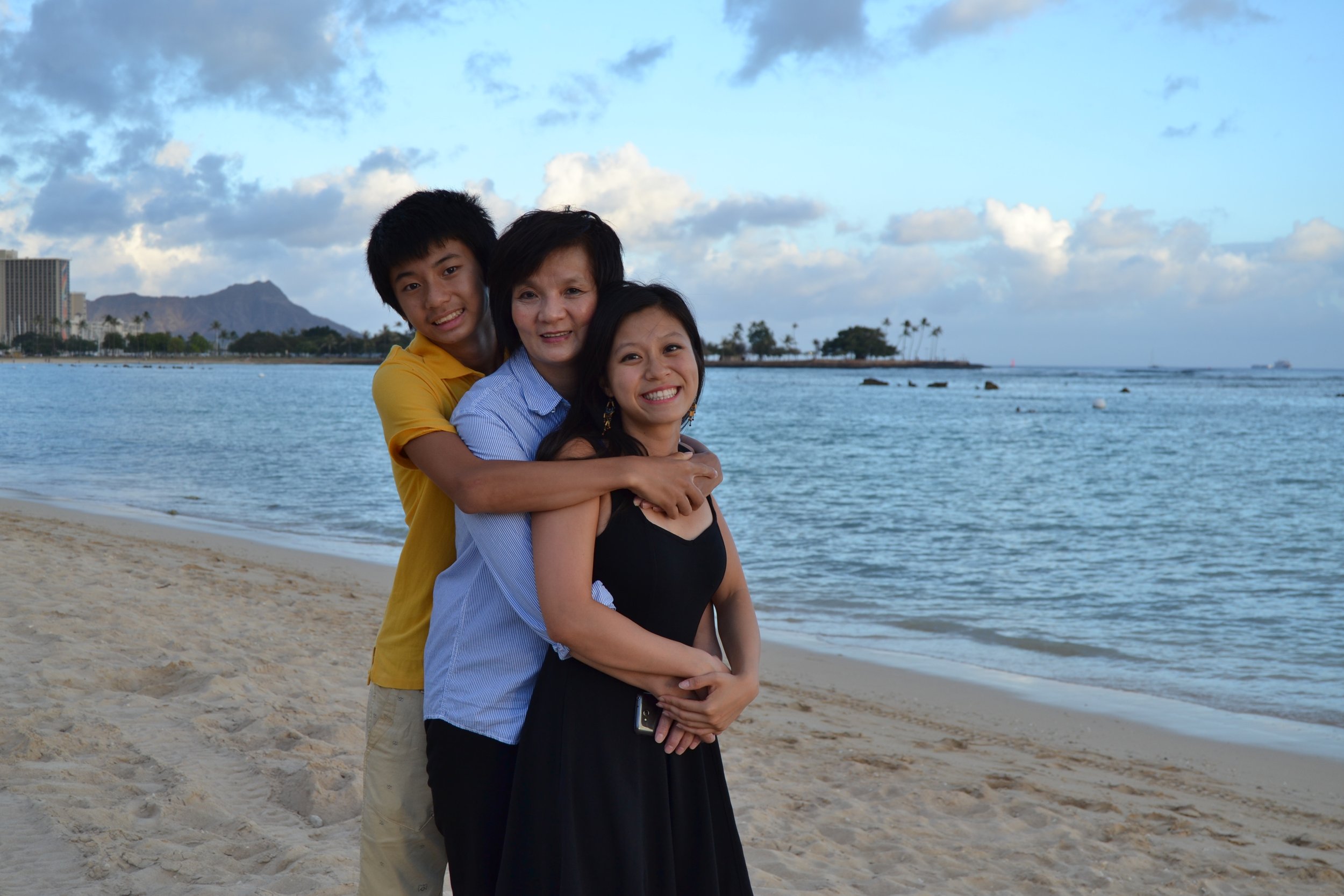 three family members embracing one another on the sands of Honlulu's Magic Island white sand beach in the summer with smiles on their faces