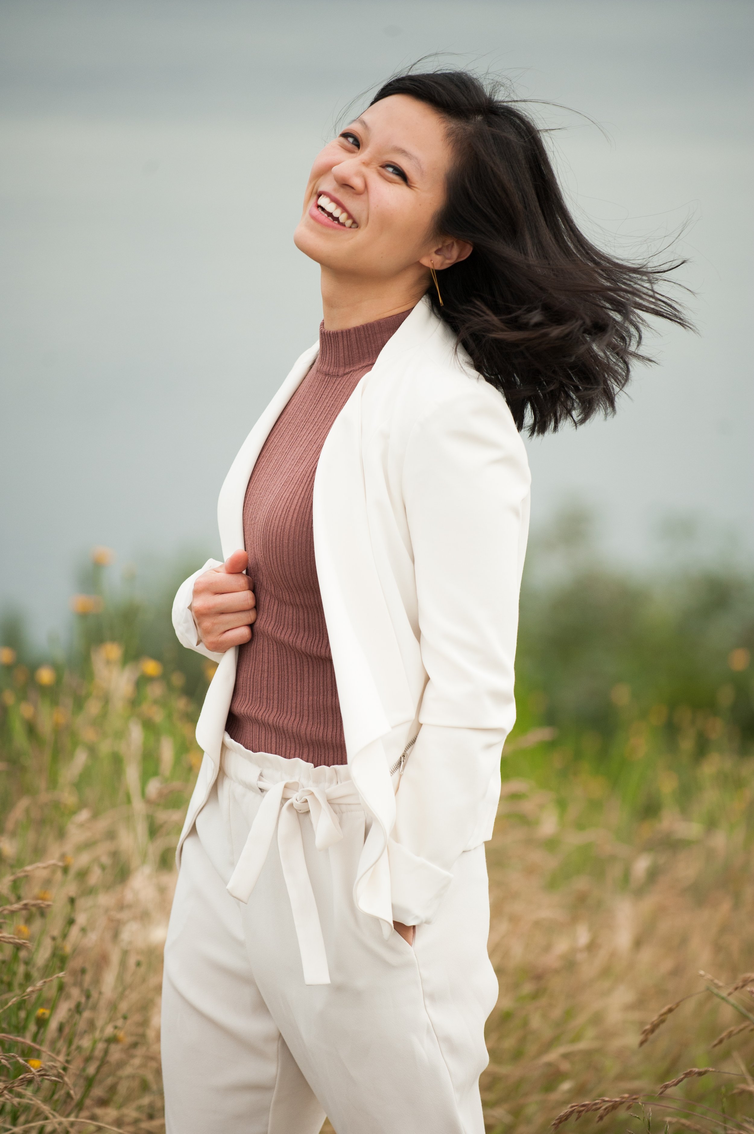 Founder of Authenticity Unearthed laughing freely with head tilted back, hair wind-blown, wearing white blazer over mauve ribbed top and white pants in an overcast wildflower field