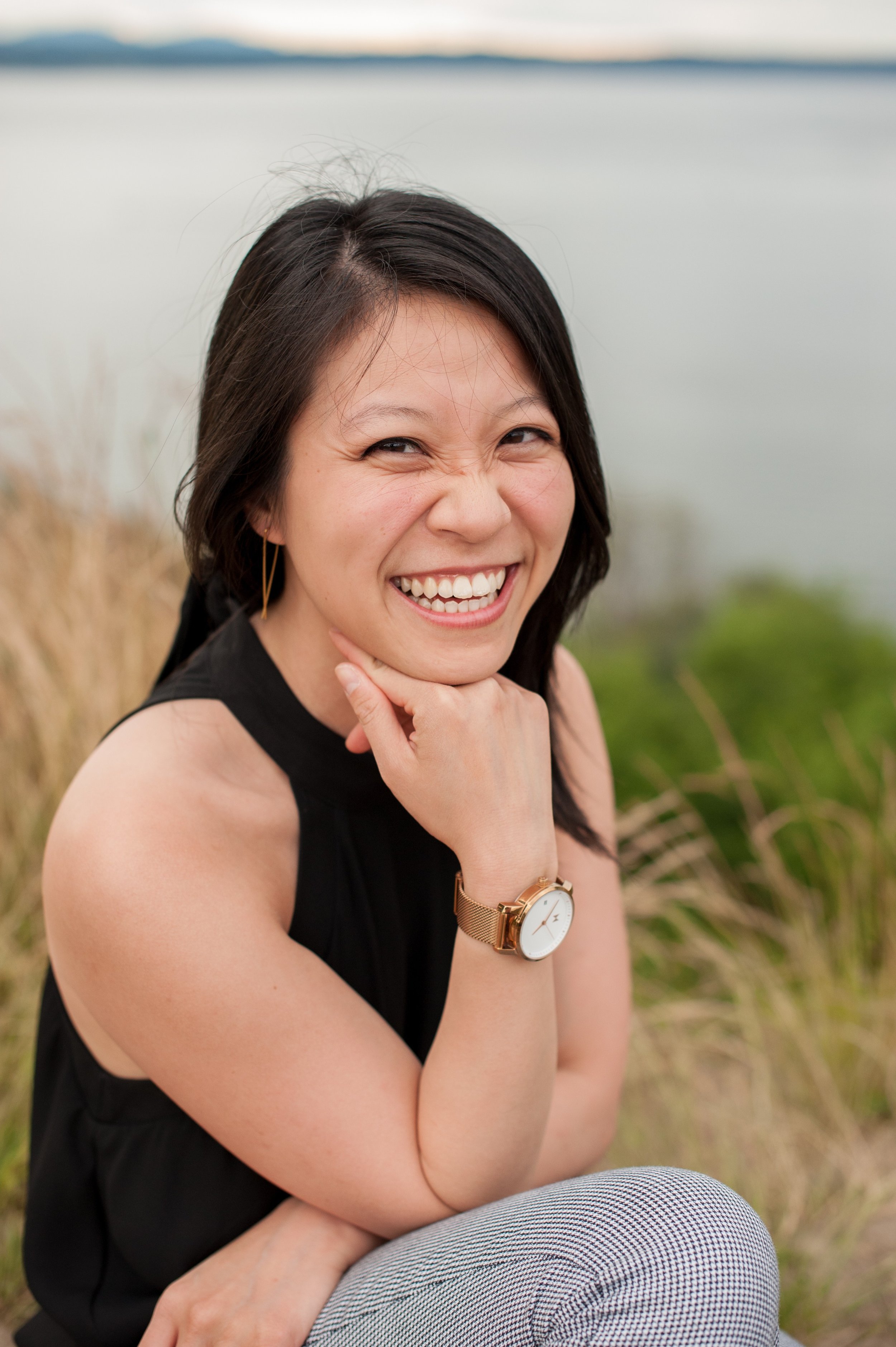 A smiling woman with short black hair, wearing a black sleeveless top, gold watch, and gold earrings, sitting outdoors near a body of water with plants in the background.
