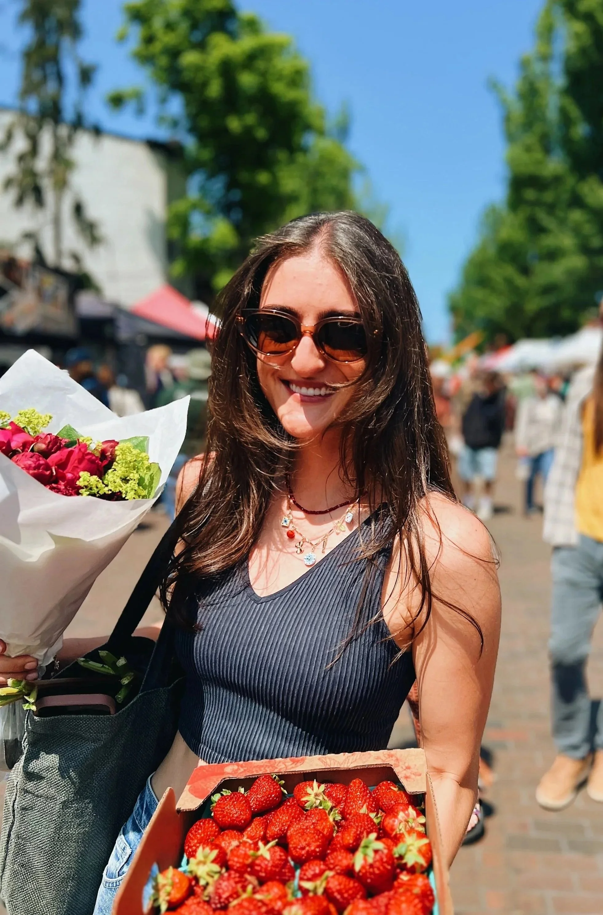 A woman with long brown hair, wearing sunglasses, a striped tank top, and necklaces, is smiling and holding a box of strawberries. She also has a bouquet of flowers. The background shows an outdoor market with trees, tents, and people.