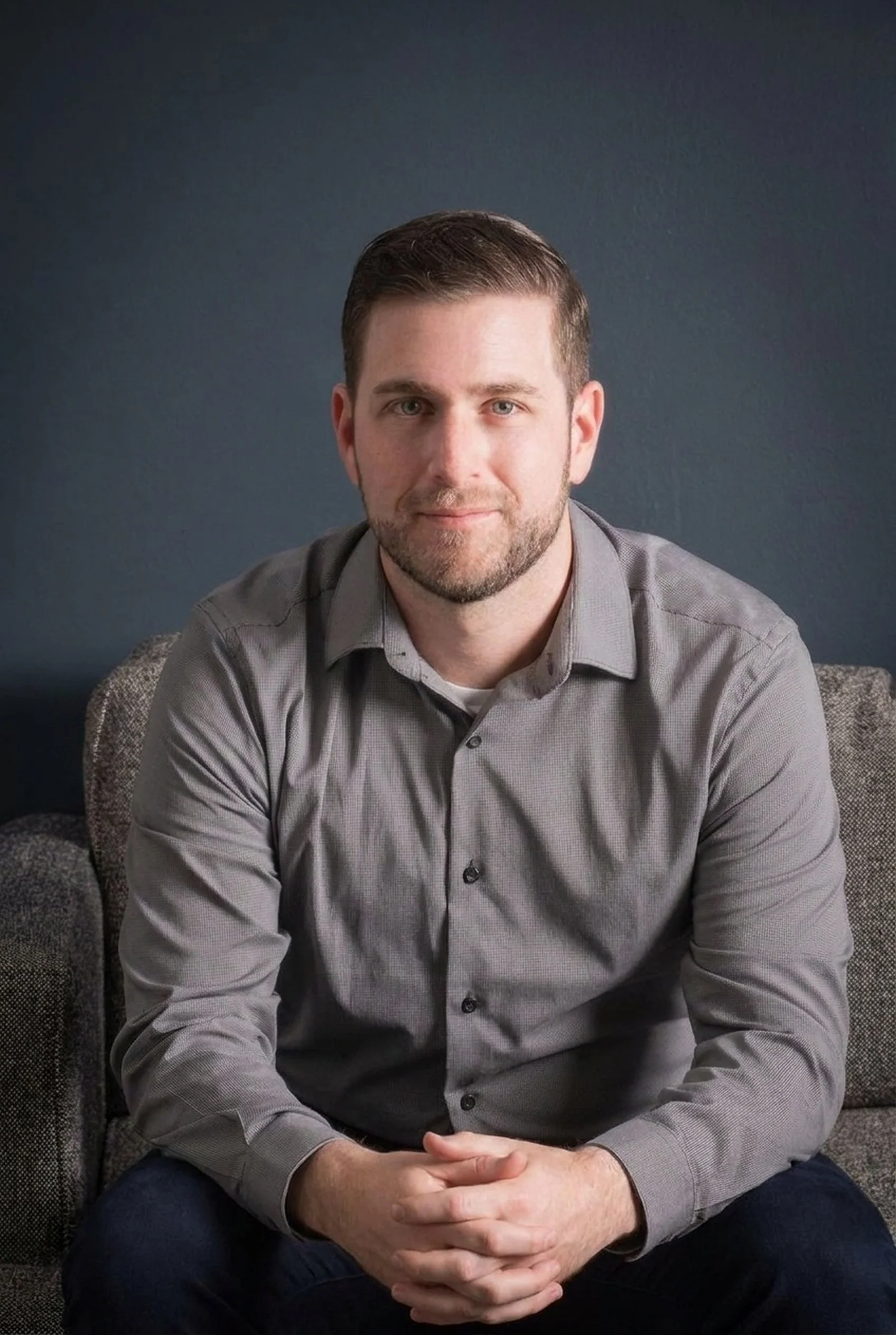 Portrait of a young man with short brown hair and a beard, wearing a gray button-up shirt, sitting on a chair with hands clasped, against a dark background.
