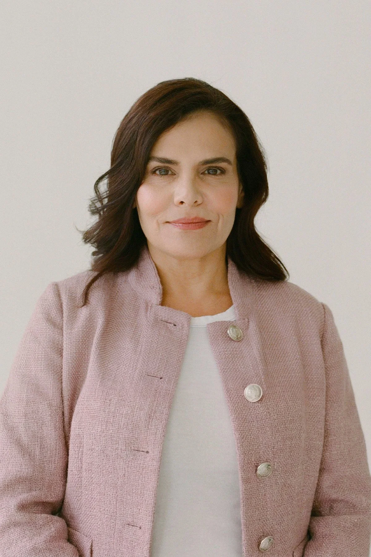 A woman with dark brown, wavy hair, wearing a pink textured blazer with large silver buttons, and a white top, standing against a plain light background.