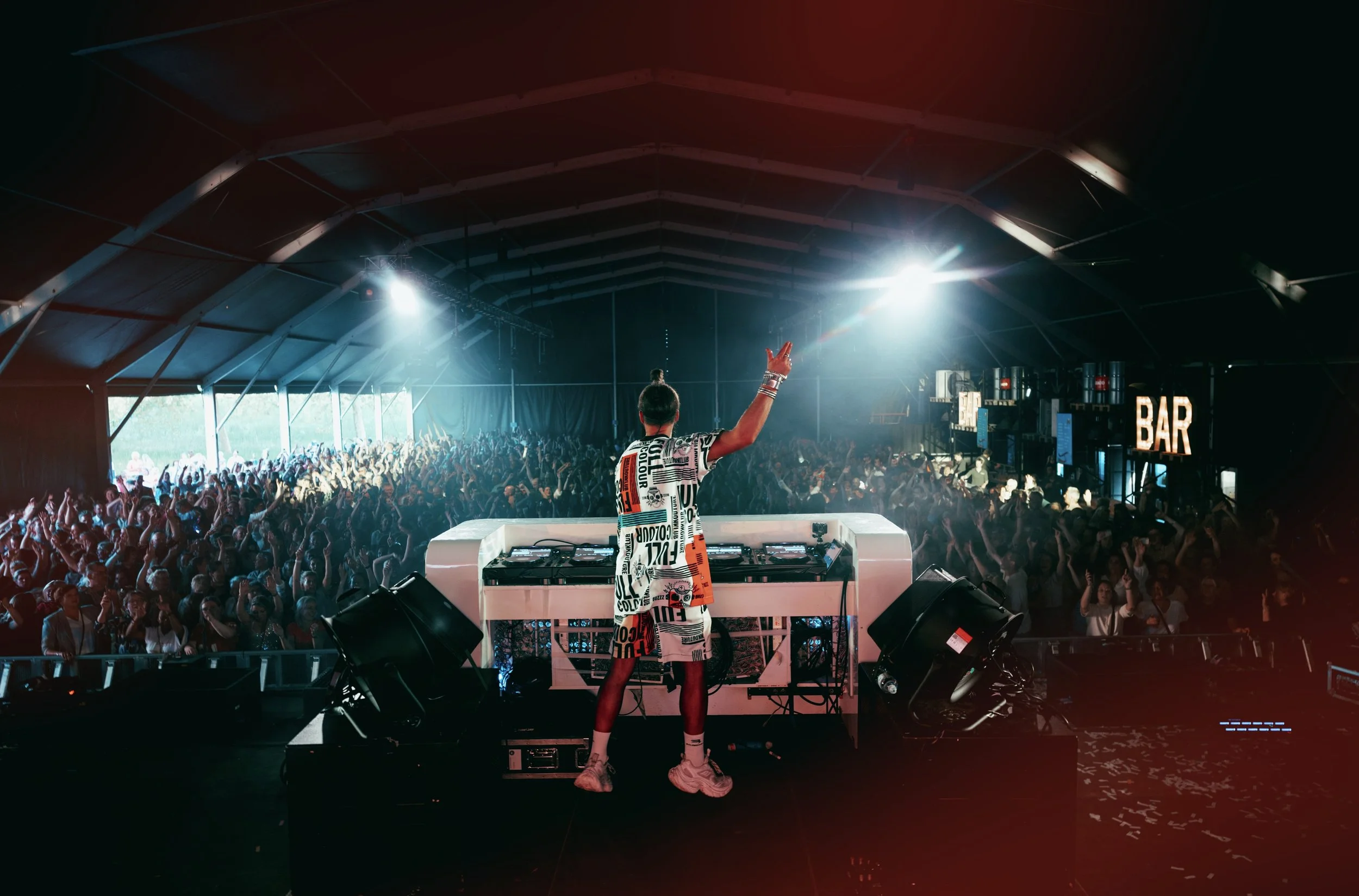 DJ performing at a music event under a large tent with a crowd dancing and enjoying the show. Bright stage lights illuminate the scene, and a bar sign is visible in the background.