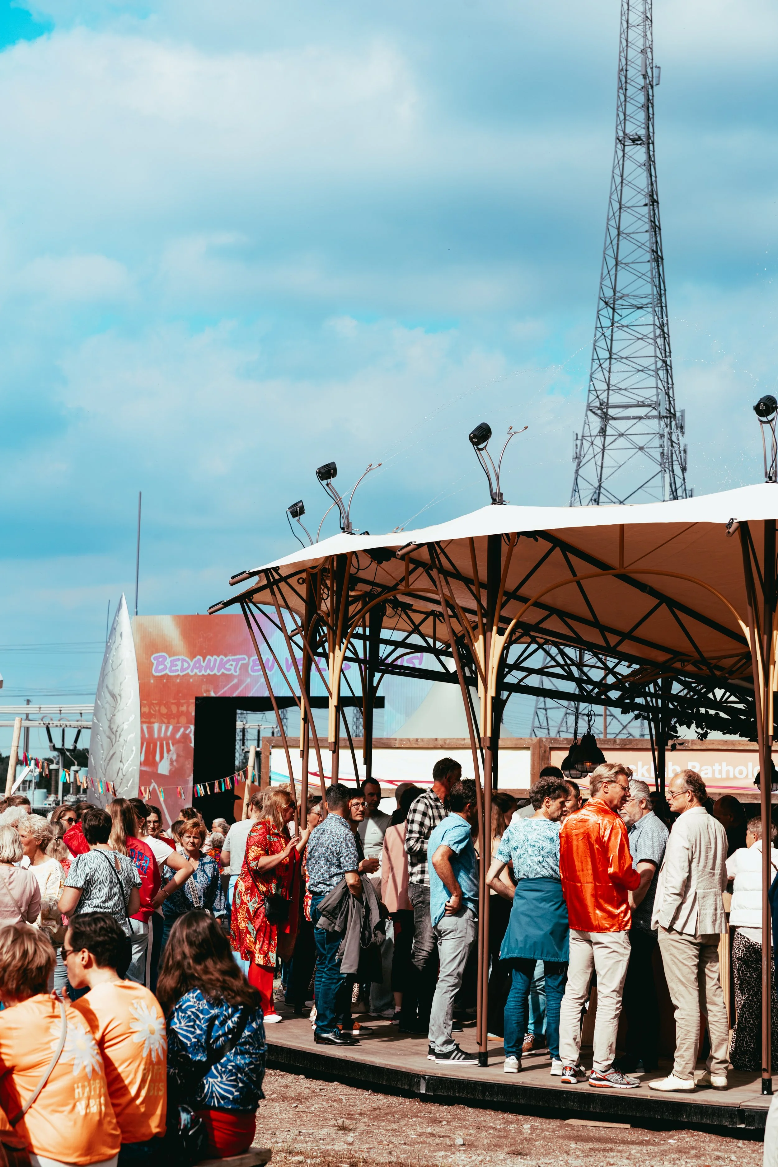 Crowd of people gathered under a large tent at an outdoor event on a sunny day with a cloudy sky in the background.