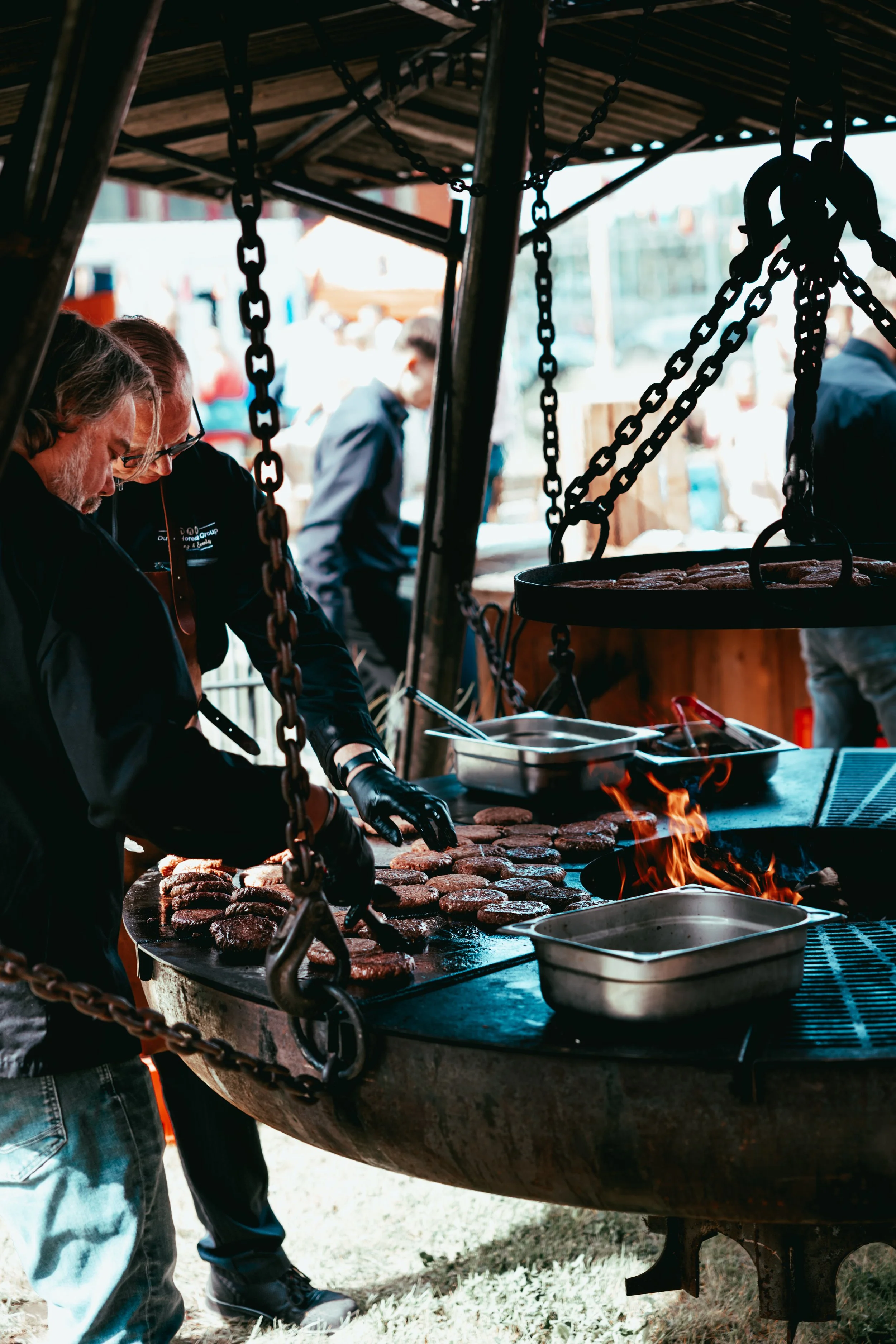 People cooking burgers on a large outdoor grill at a fair or festival.