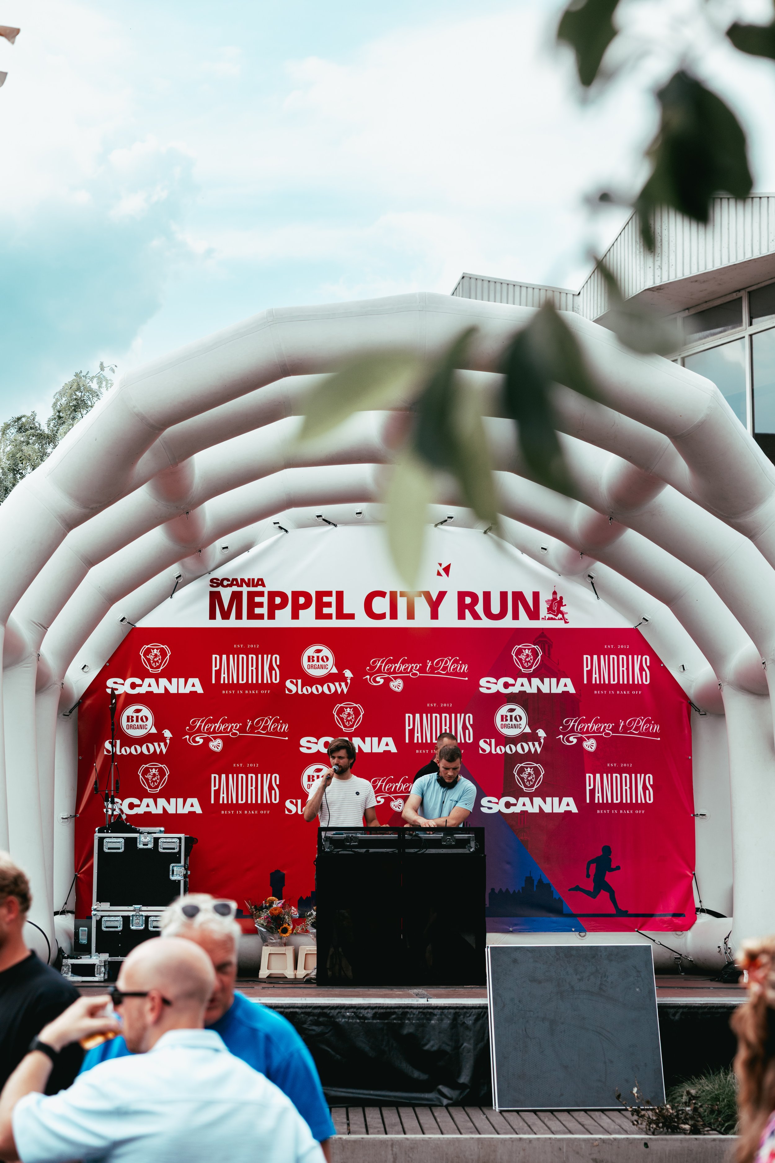 Two men are performing on a stage at the Meppel City Run event. The stage has a red backdrop with event sponsors' logos and a silhouette of a person running. People are in the audience, some taking photos.