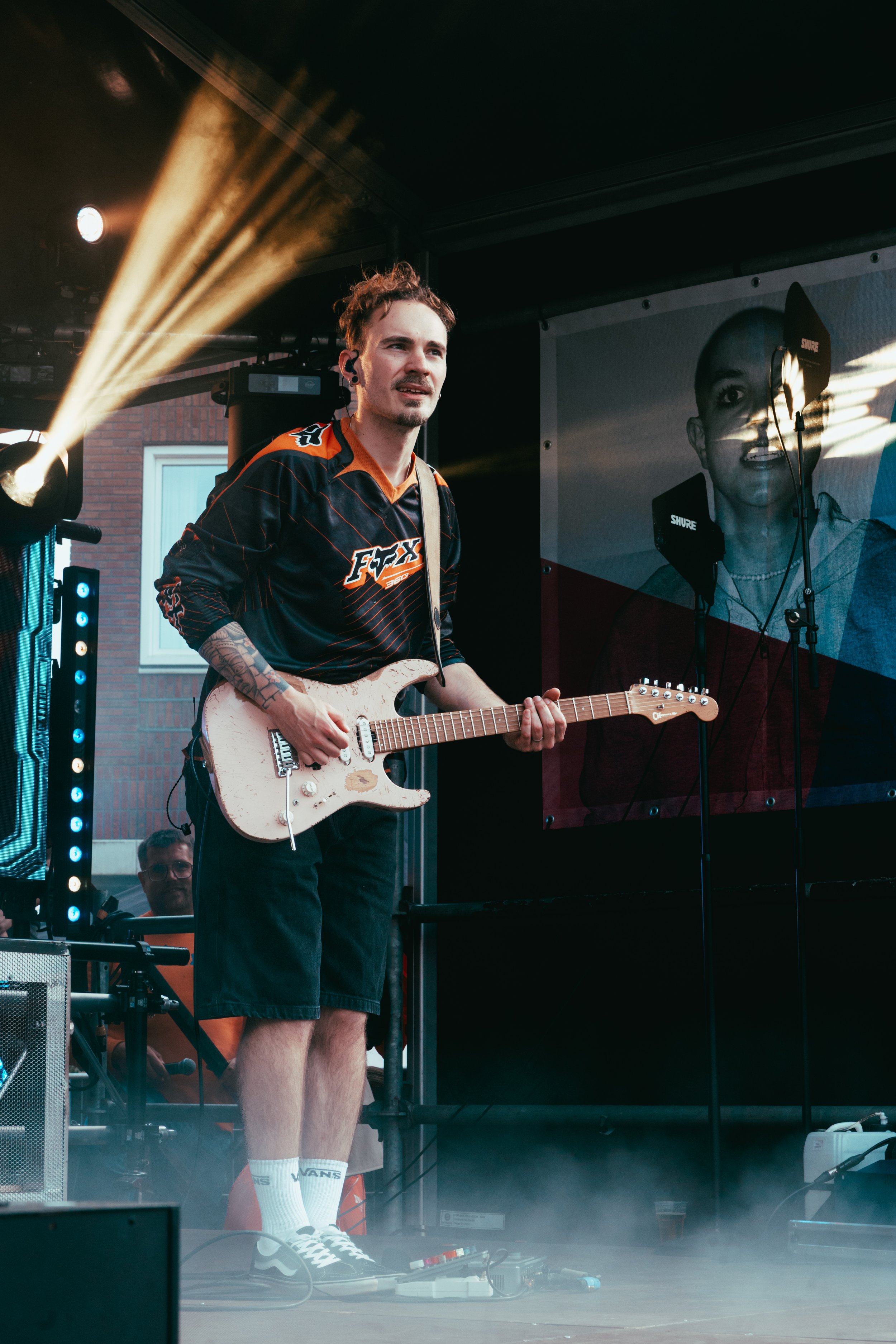 A young man with a guitar performing on stage at an outdoor music event, wearing a black and orange sports jersey, shorts, and Vans socks.