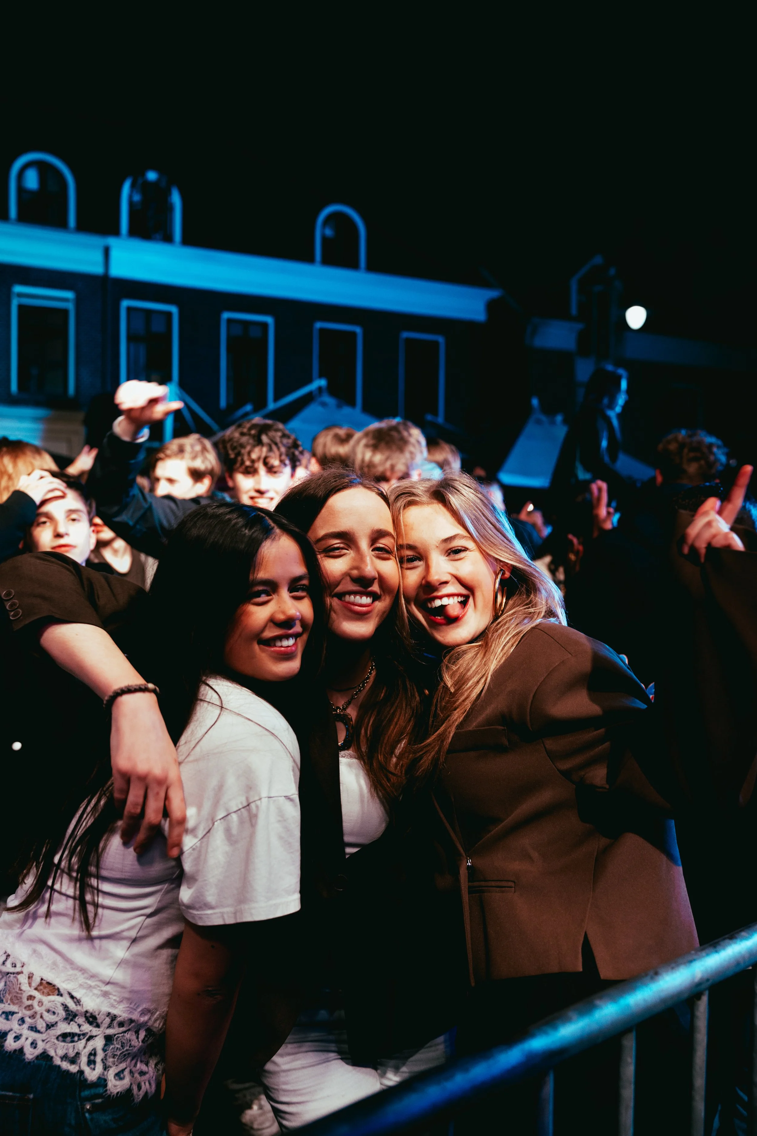 Group of smiling young women embracing each other at a concert or party, with a crowd of people in the background and nighttime city buildings visible.