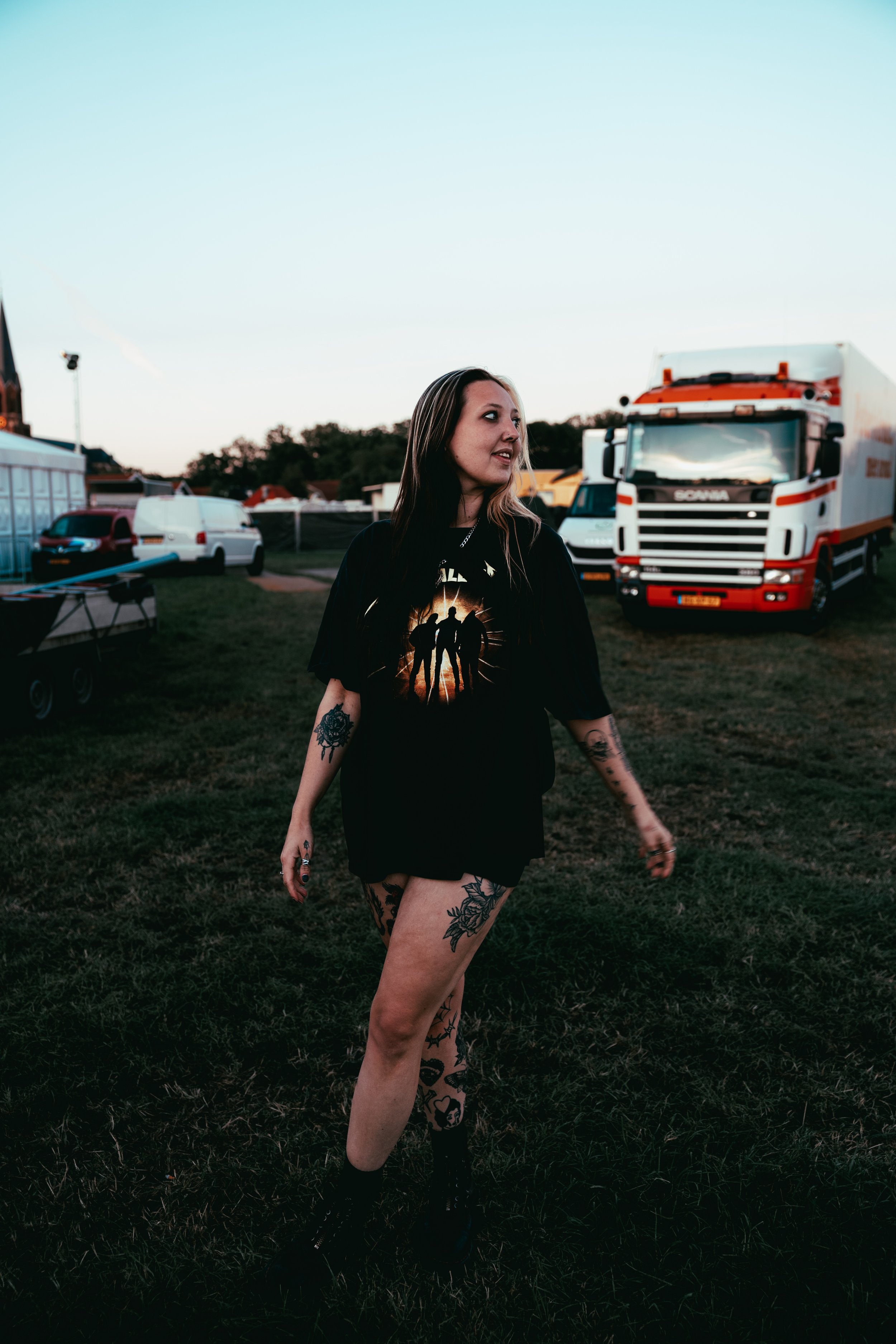 A young woman with tattoos on her arms and legs standing outdoors during sunset, wearing a black graphic T-shirt, black shorts, and black boots. Behind her are police cars, trucks, and tents in an open field.