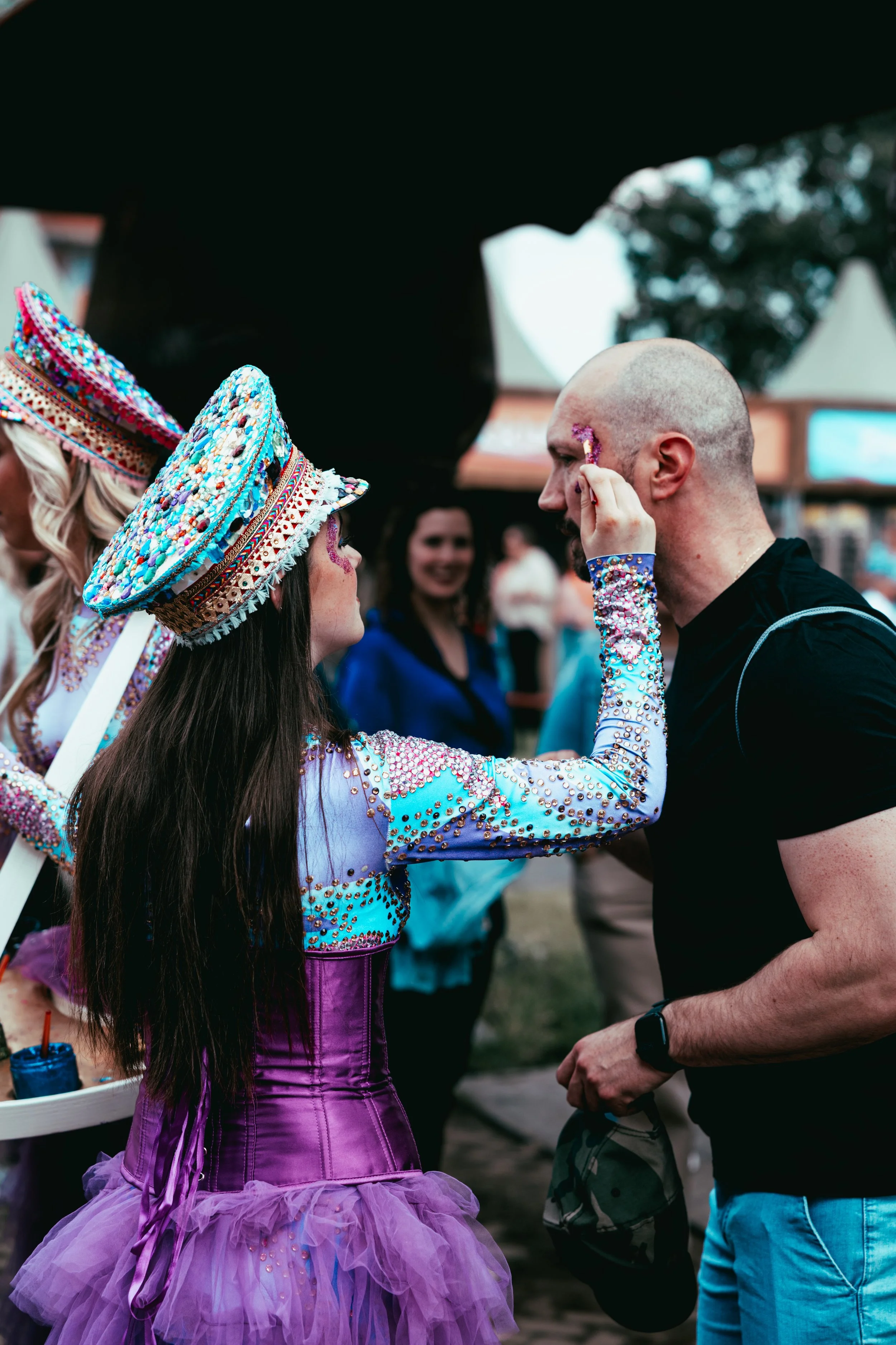 A woman in a colorful, beaded costume and a wide-brimmed, decorated hat decorates a man's face with glitter at an outdoor event. She has long dark hair, and the man has a shaved head and wears a black shirt and camouflage pants. Other people are in t