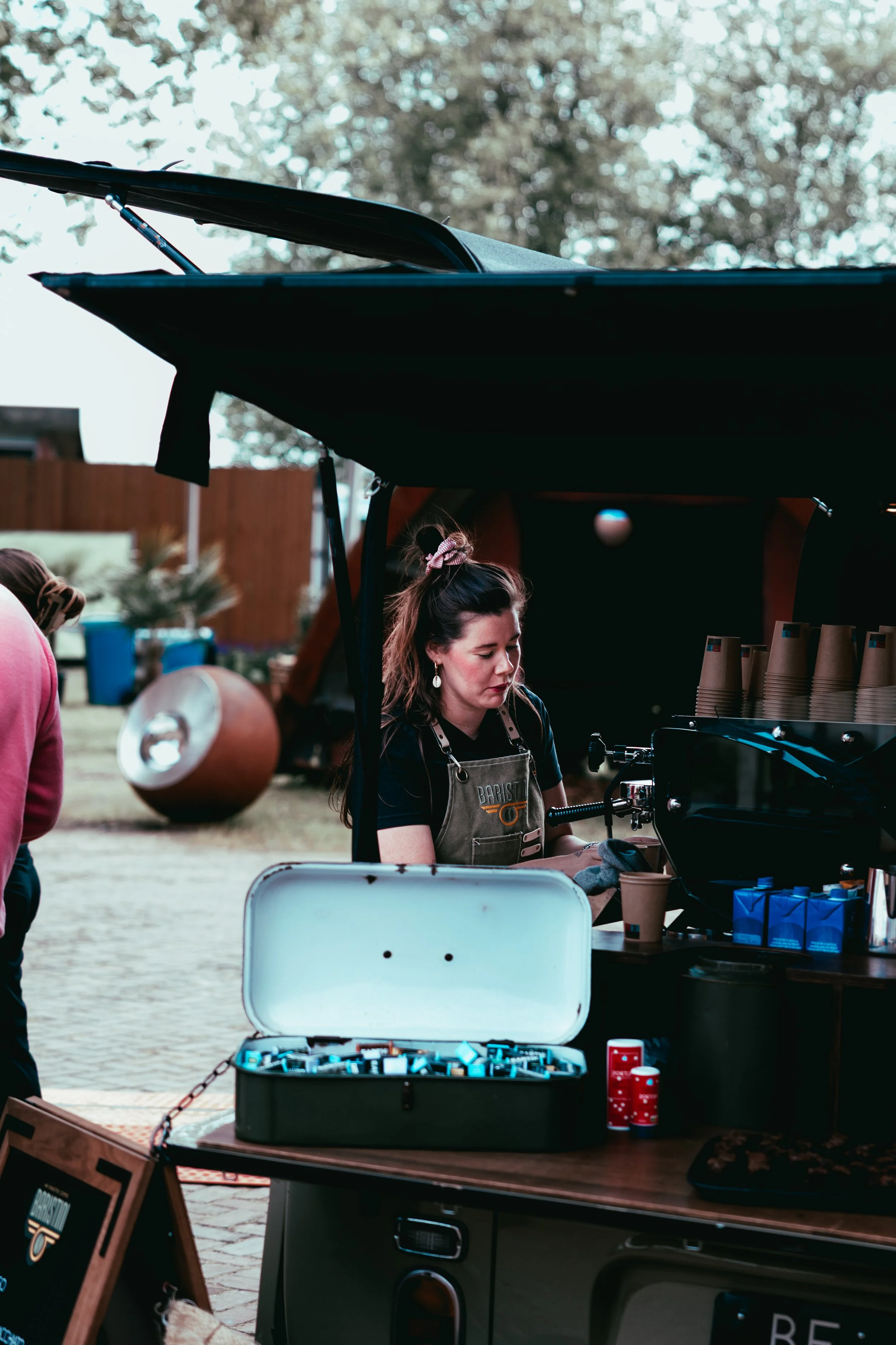 A woman operating an espresso machine at an outdoor coffee stand, with disposable coffee cups, a small table with a box of blue pods, and Christmas-themed red cups nearby.