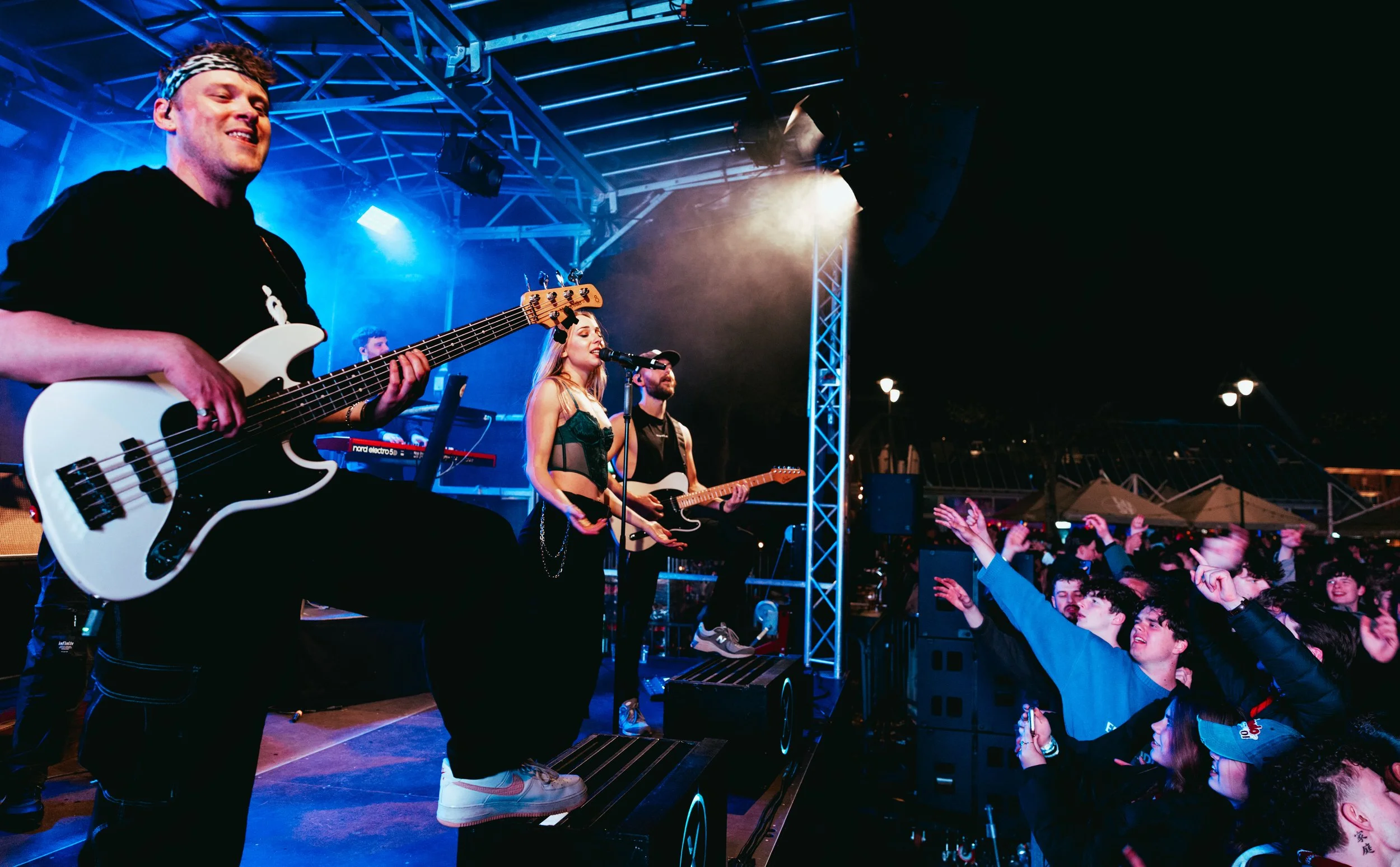 Musicians performing live on stage at night with a cheering crowd in front, illuminated by stage lighting and metal framework.