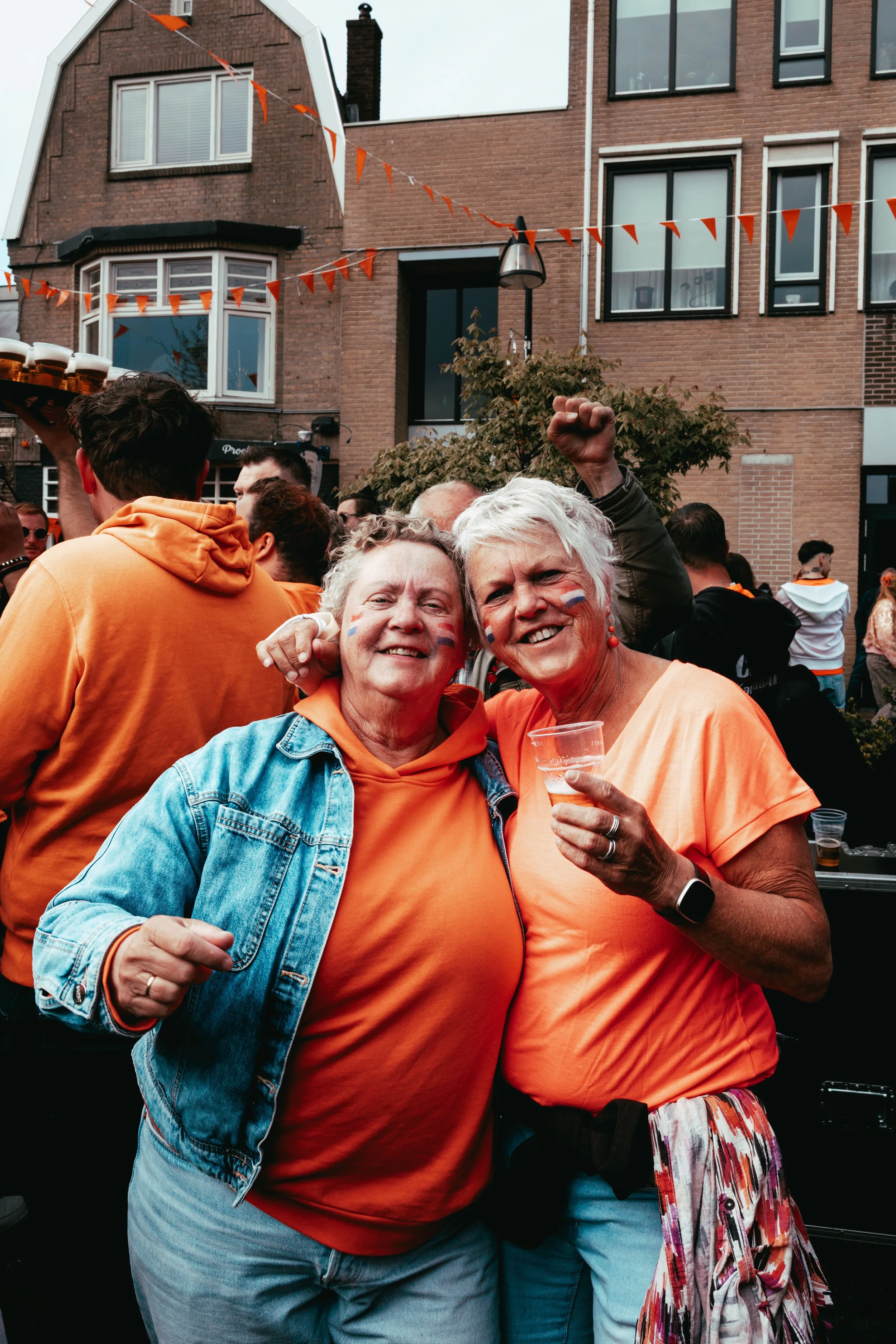Two older women with painted faces smiling and holding drinks at an outdoor gathering, wearing orange shirts. They are surrounded by other people and festive decorations, with a brick building in the background.