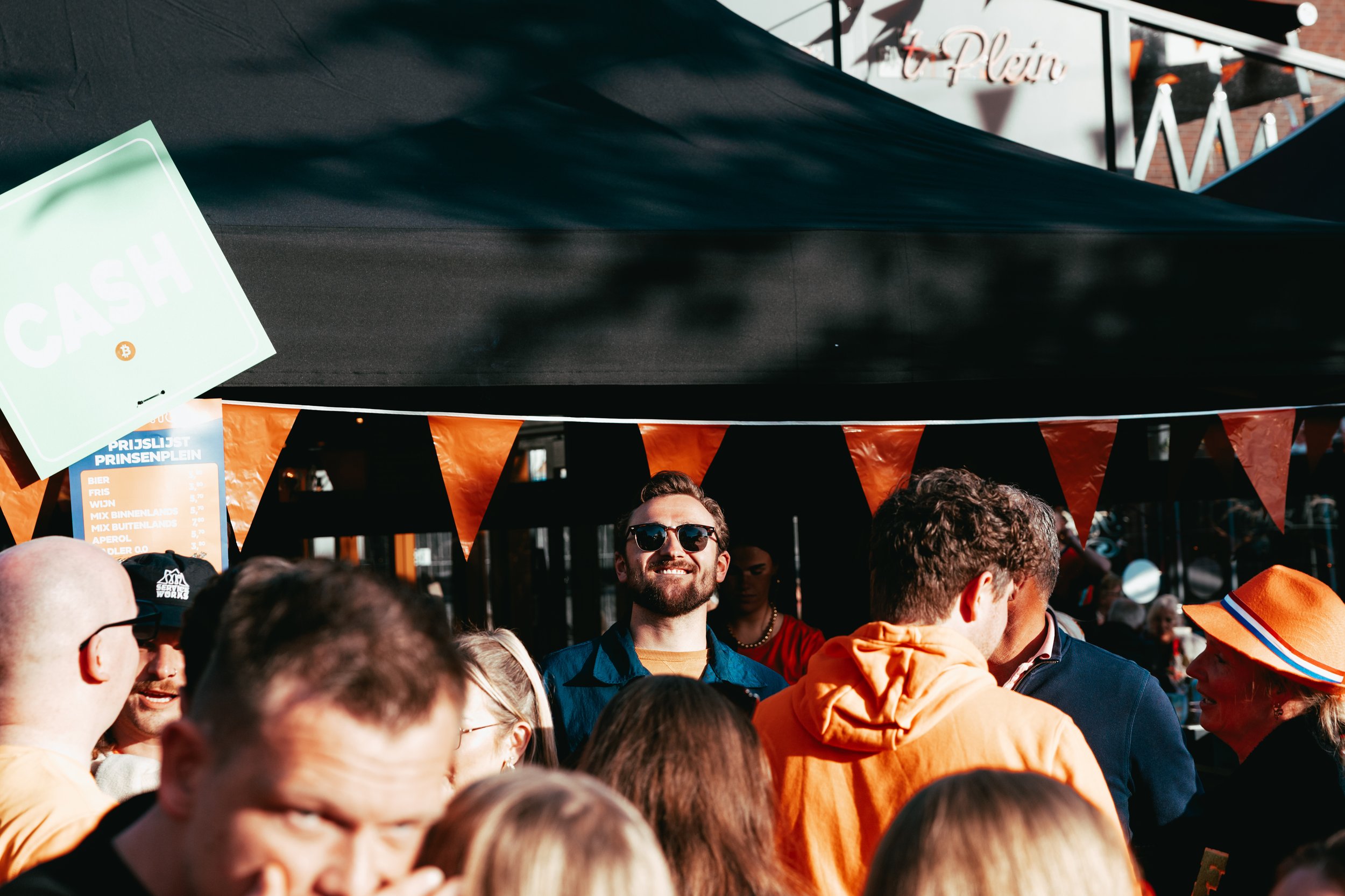 Crowd of people gathered outdoors on a sunny day, with some wearing orange clothing. He stands in the center wearing dark sunglasses, smiling. There are signs and a building in the background.