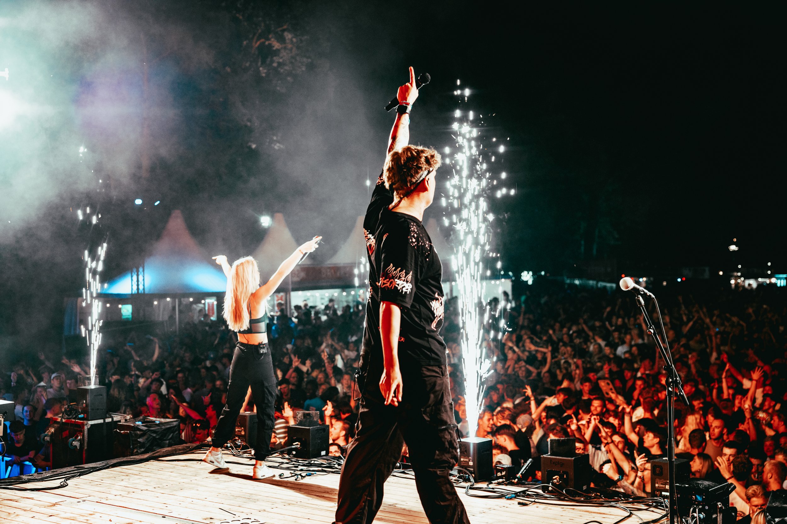 A performer on stage holding a microphone and raising their hand with one finger pointed up during a concert. There are fireworks and a large crowd in the background.