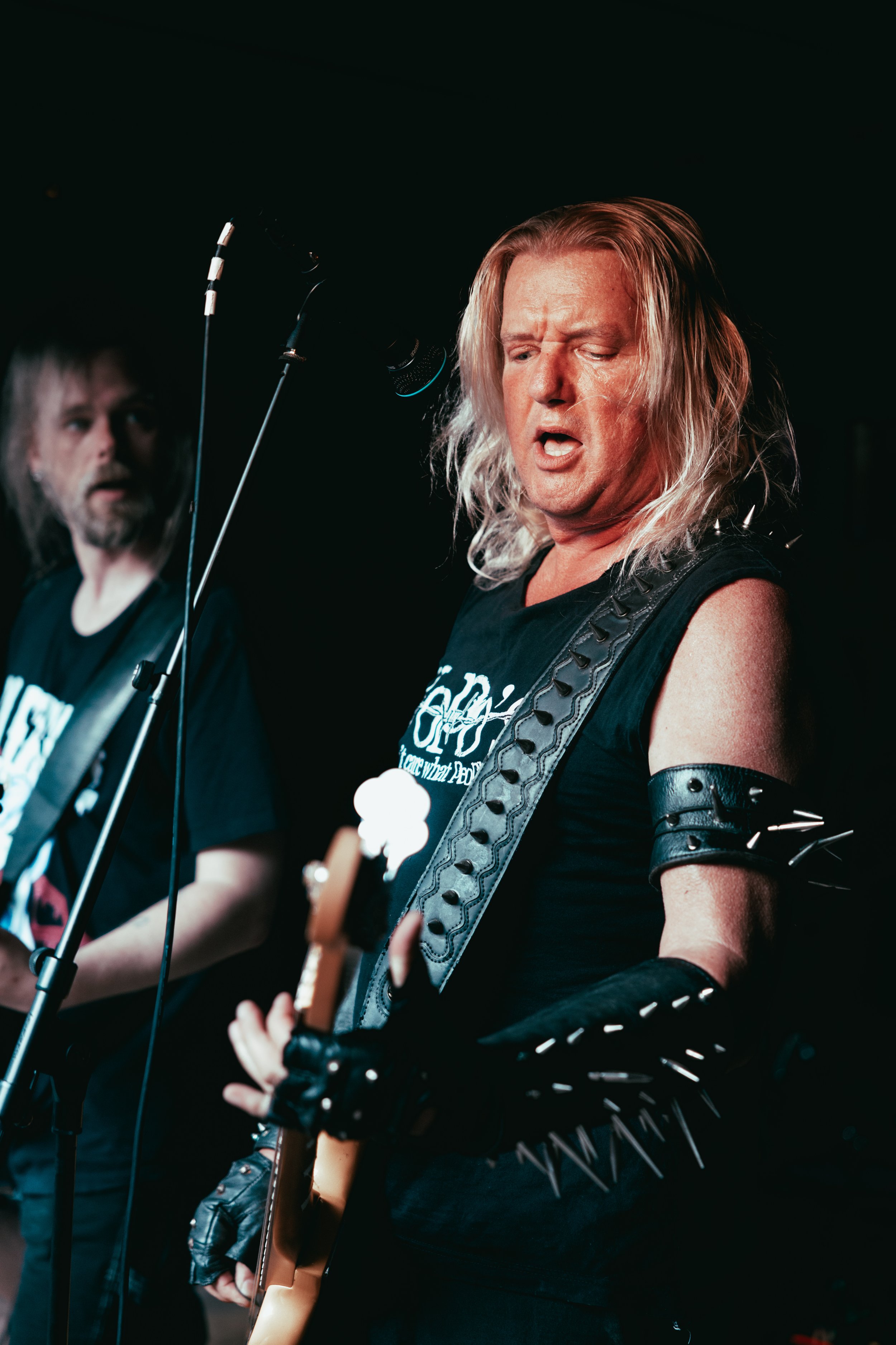 Two guitarists performing on stage, one with long blond hair, black sleeveless shirt, and black gloves with spikes playing an electric guitar, the other with long dark hair and beard, also playing guitar, in a dark setting.