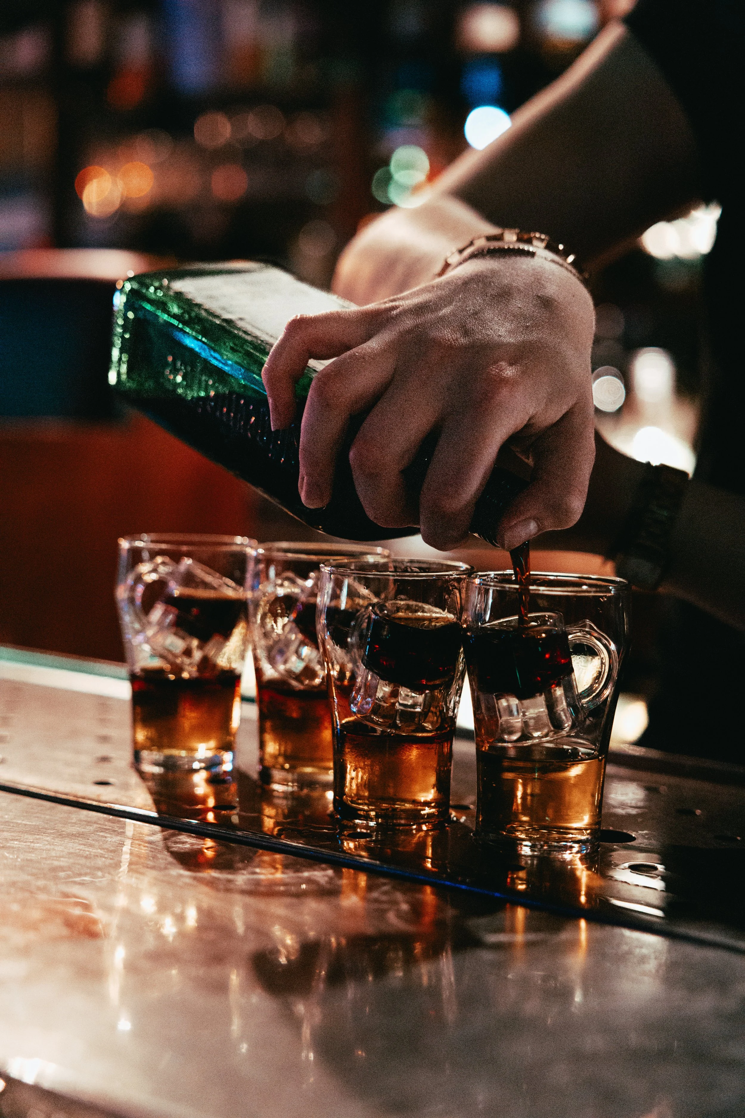 A person pouring an alcoholic beverage into shot glasses on a bar counter at night, with blurred city lights in the background.