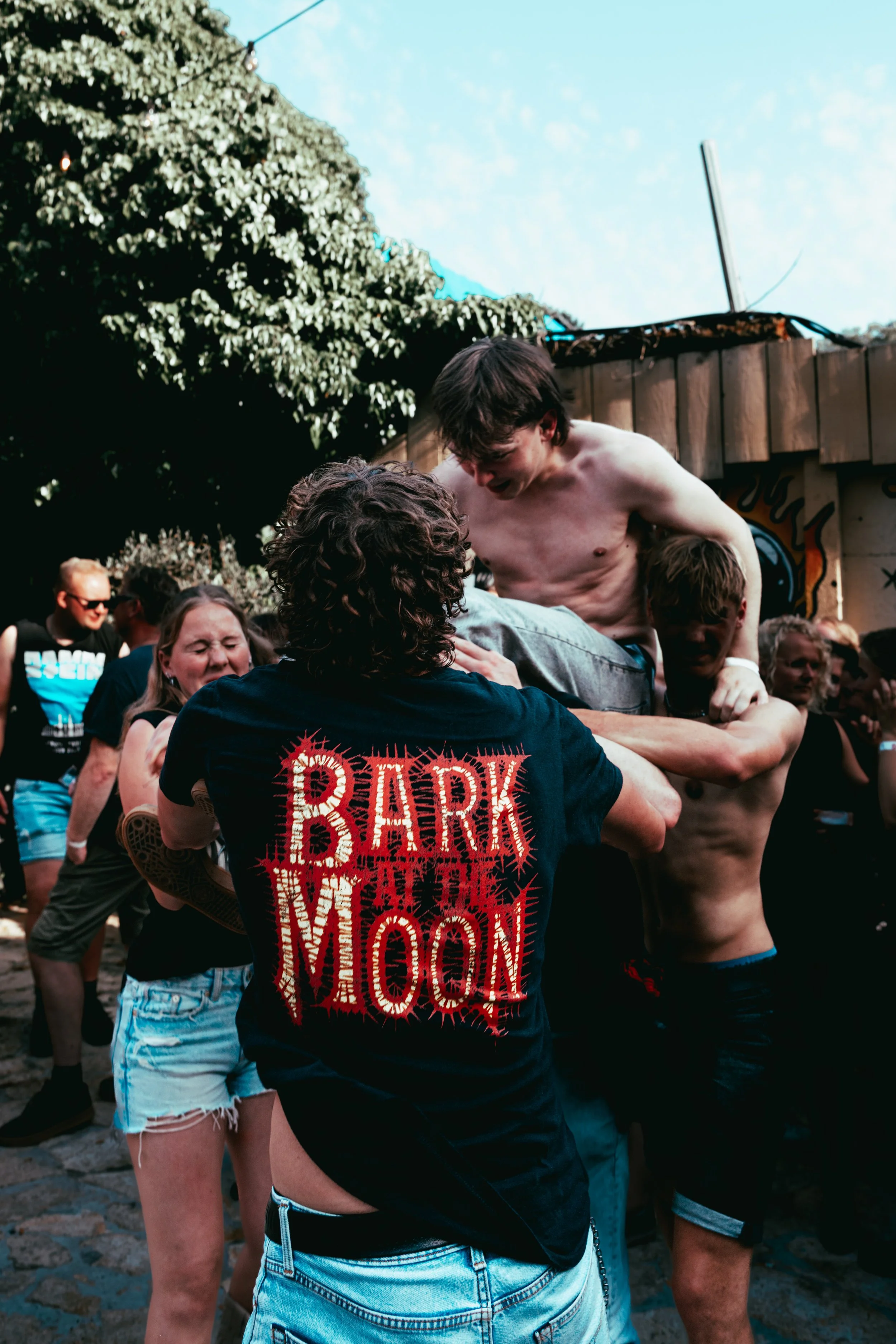 A group of people outdoors, with a shirtless man being lifted or crowd-surfed by others. The man has dark hair and is smiling. Several other people are visible in the background, some smiling and enjoying the event, with trees and a wooden structure 