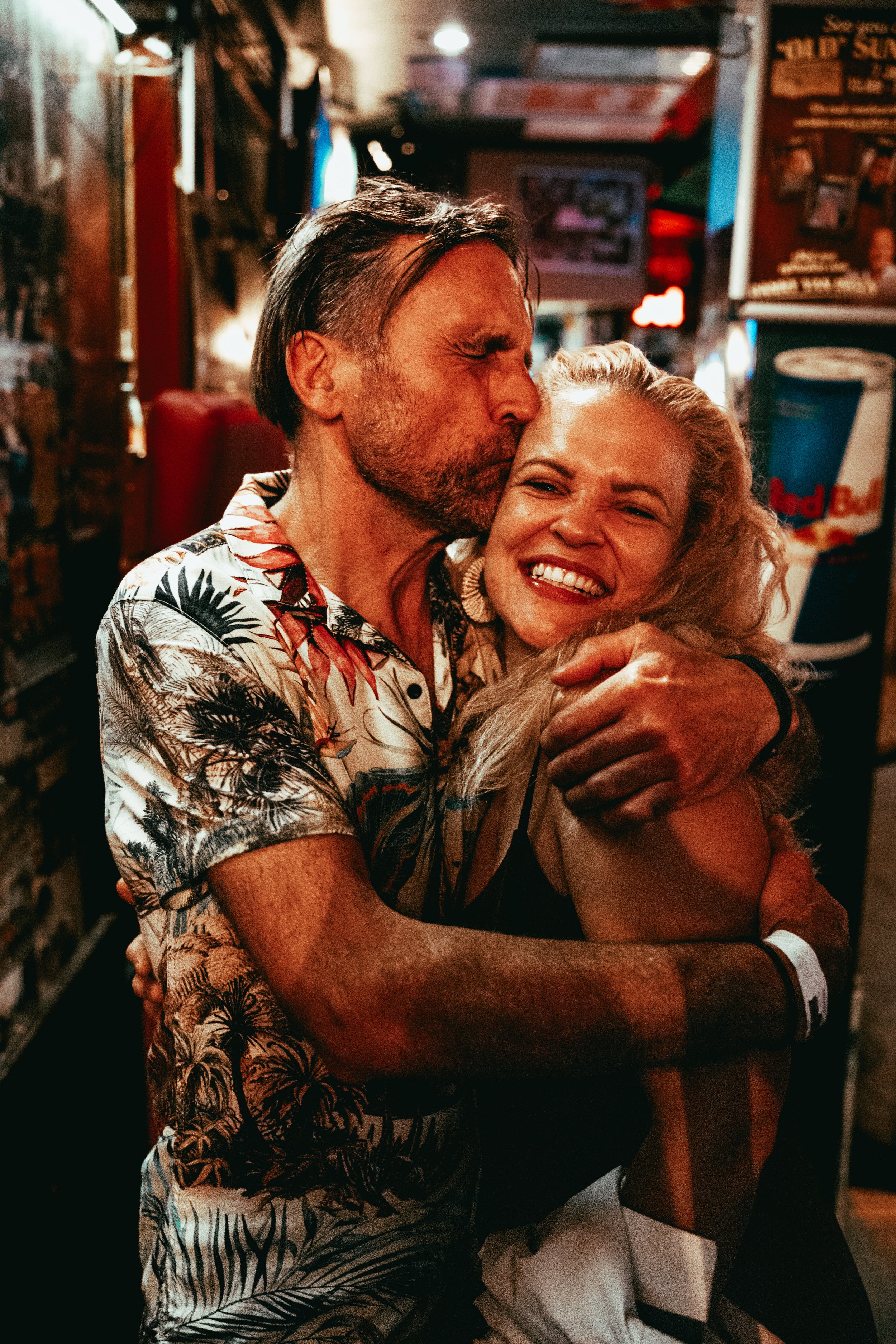 A man and a woman hugging and smiling in a dimly lit bar or restaurant. The man is kissing the woman on the forehead while she smiles happily. The background features posters and a Red Bull beverage dispenser.
