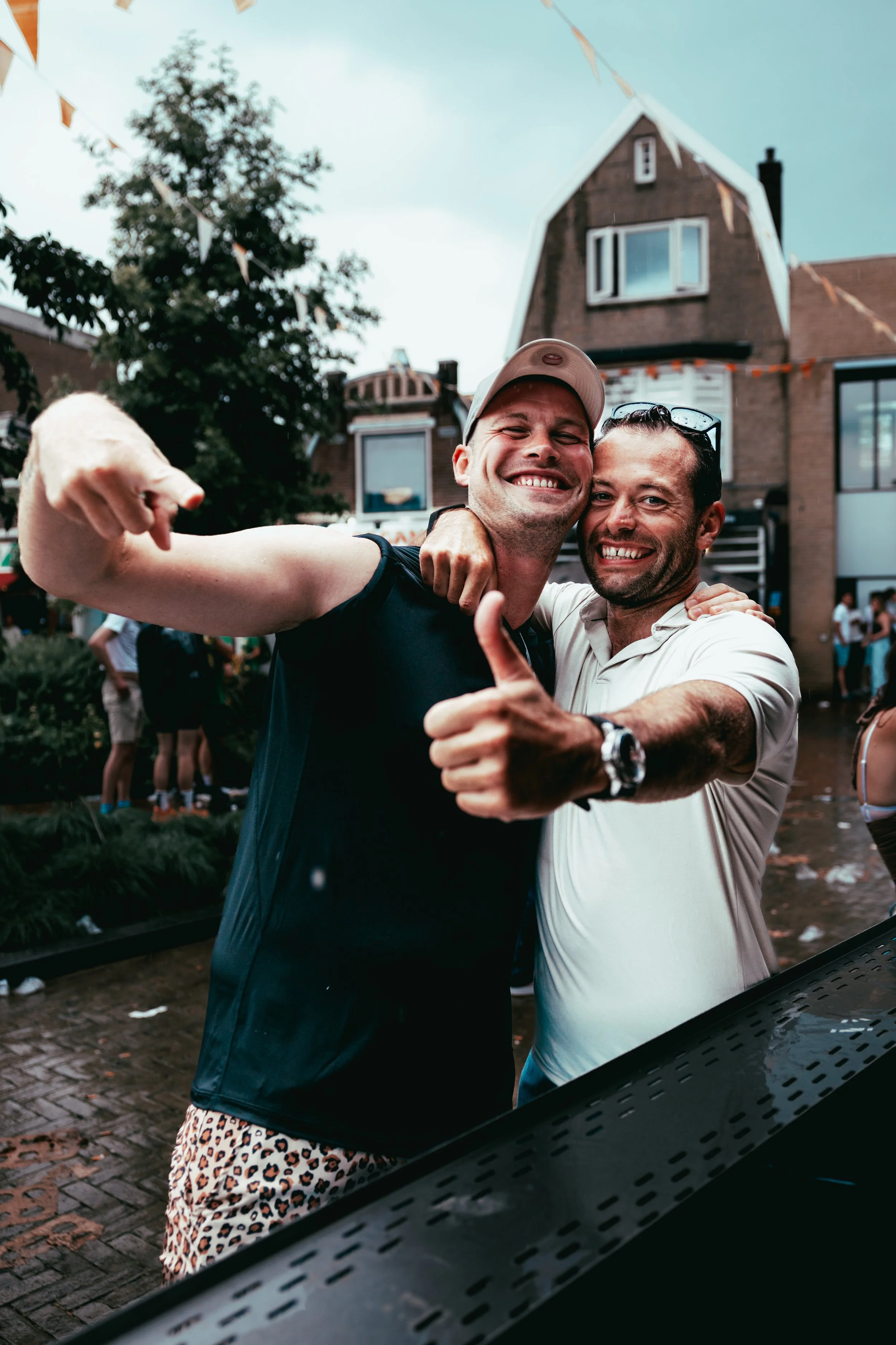 Two men smiling and embracing during an outdoor celebration in a neighborhood, with houses and decorations in the background.