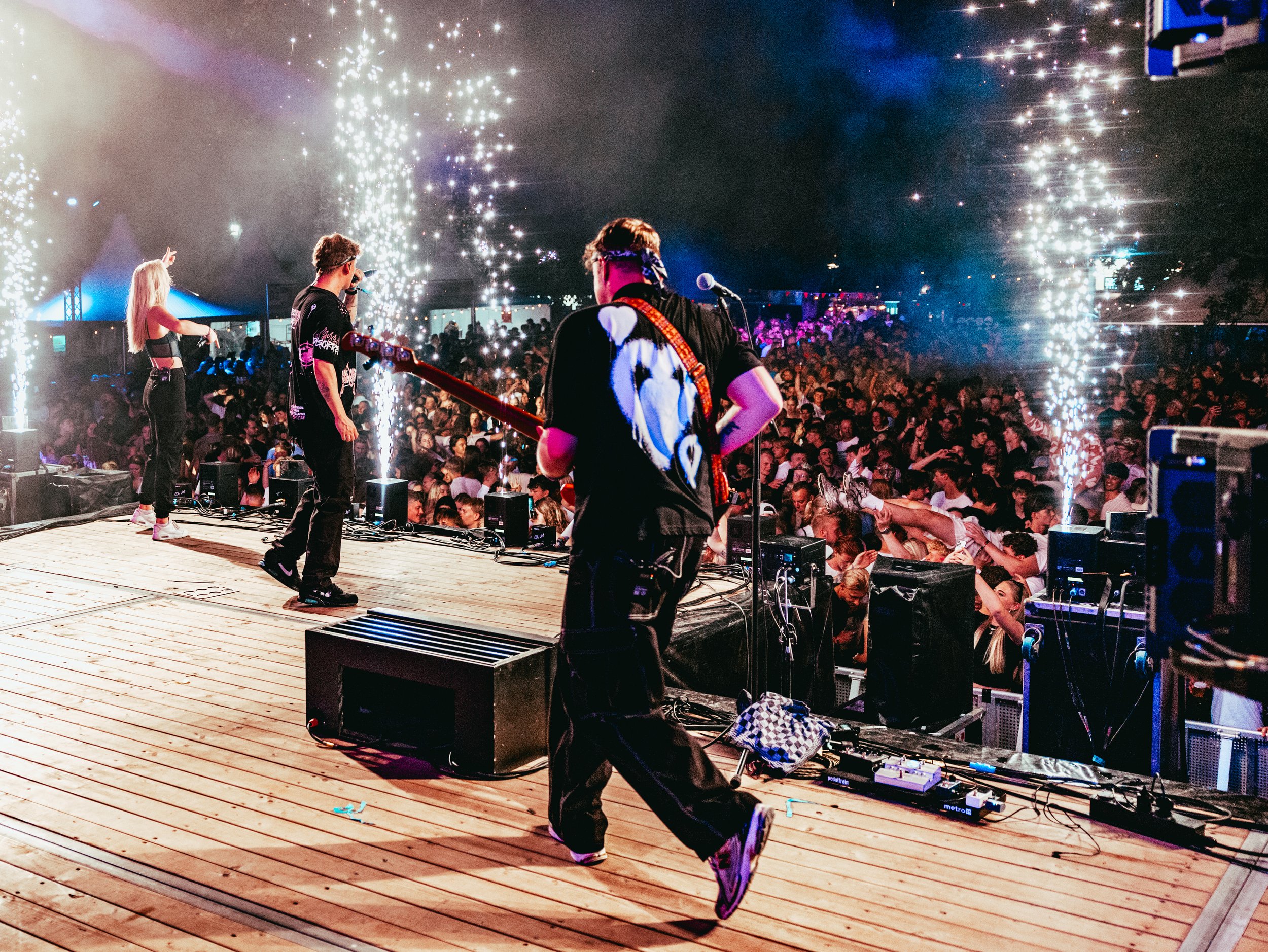 Musicians performing on a stage at a concert with a large crowd, fireworks, and colorful lights in the background.