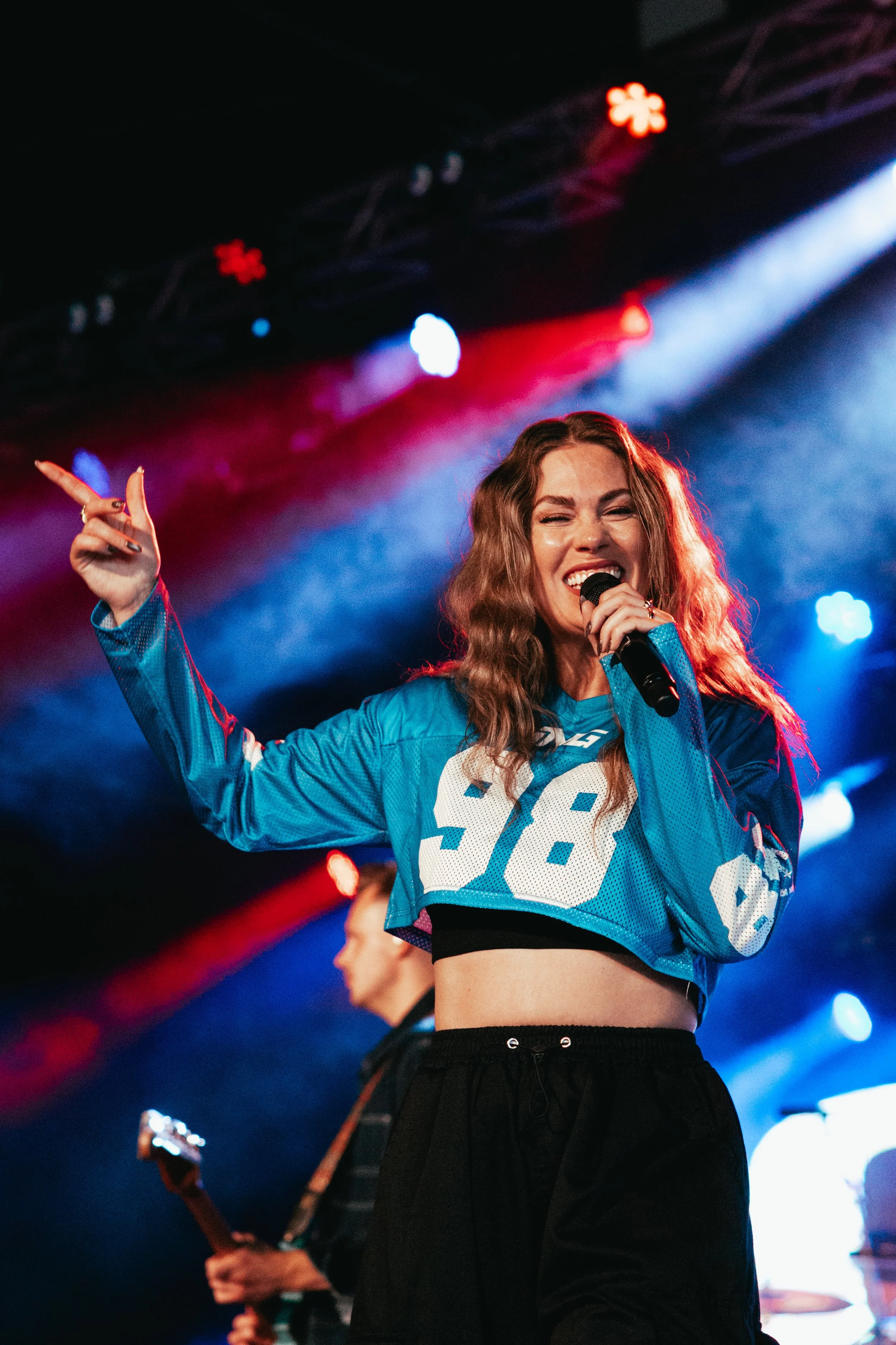 A female singer with long wavy hair singing into a microphone on stage. She is wearing a blue sports jersey with the number 28 and black pants. Bright stage lights and colored lights are visible in the background.