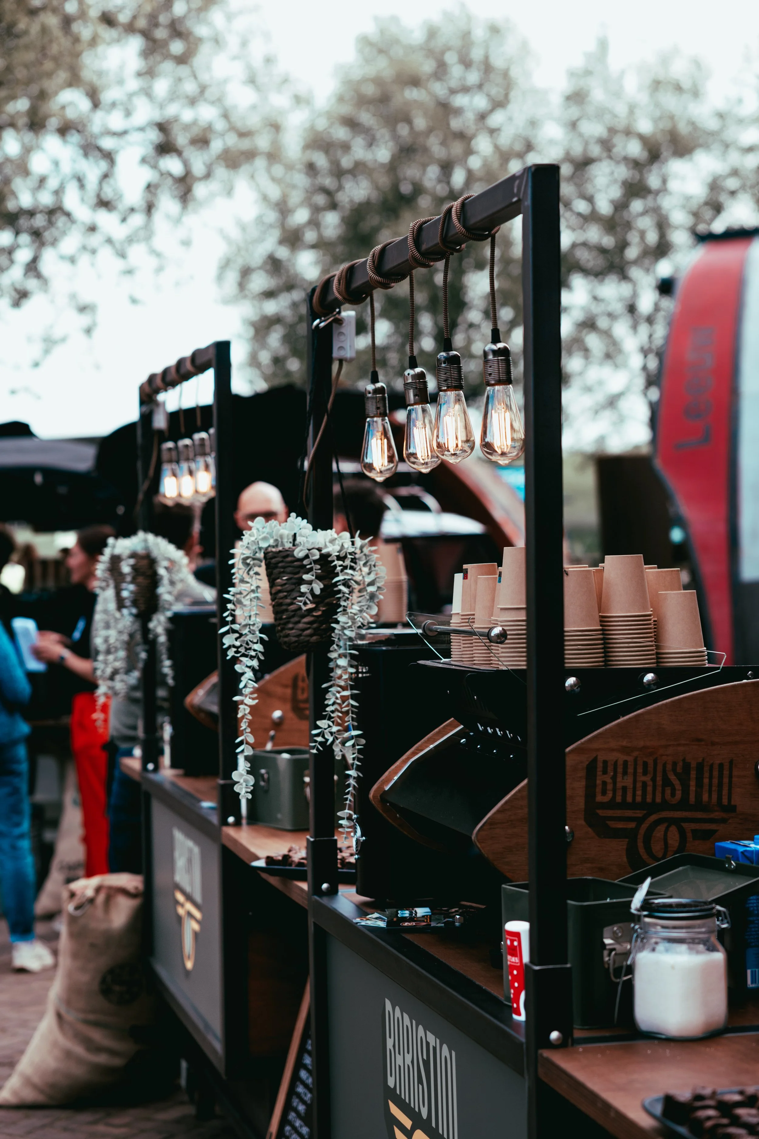 A market stall with hanging light bulbs, paper cups, and decorative items, with people in the background and trees overhead.