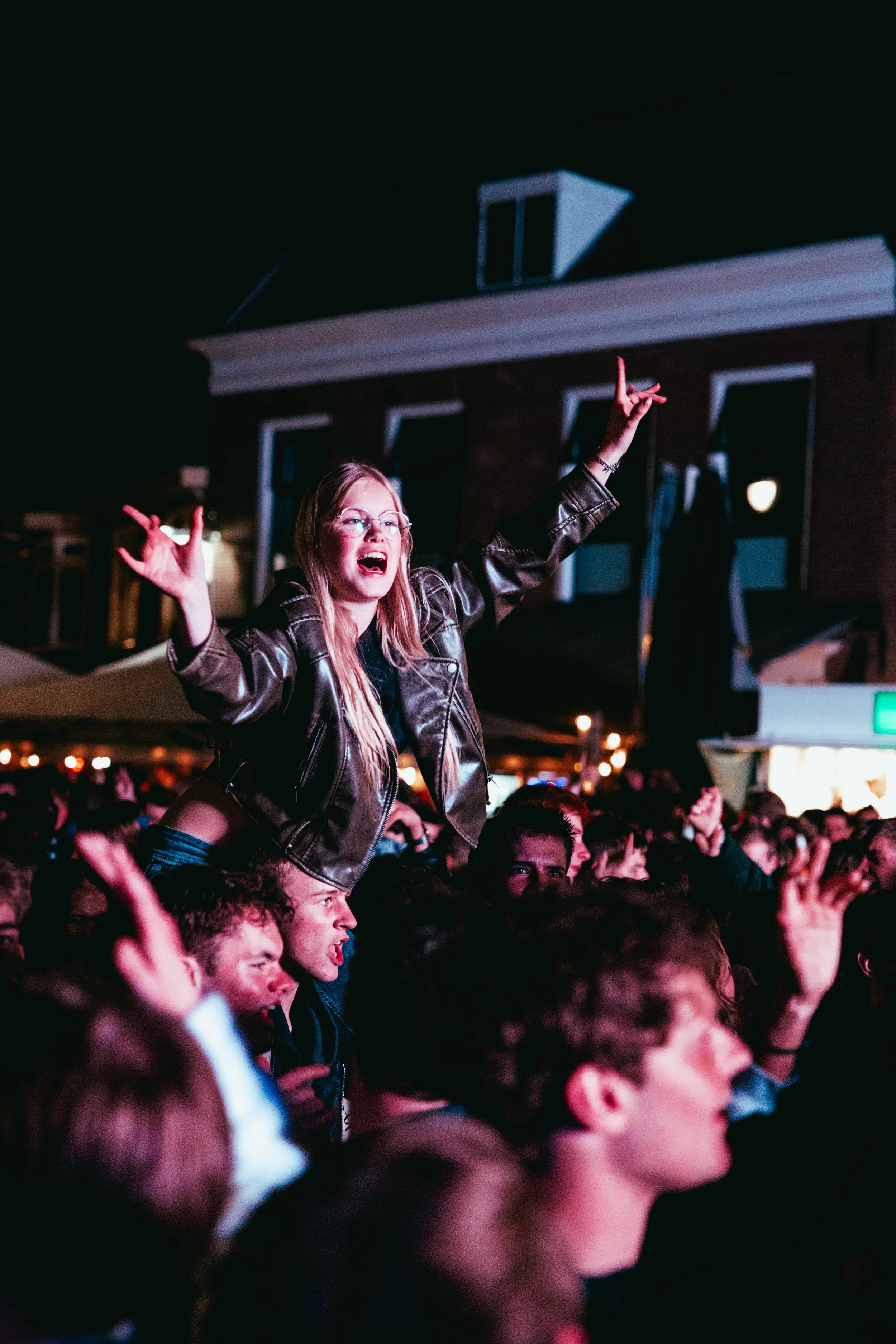 A woman standing on someone's shoulders at an outdoor concert or event at night, raising her arms with one making a rock gesture, surrounded by crowd members. BOERENMAANDAG NIJKERK.