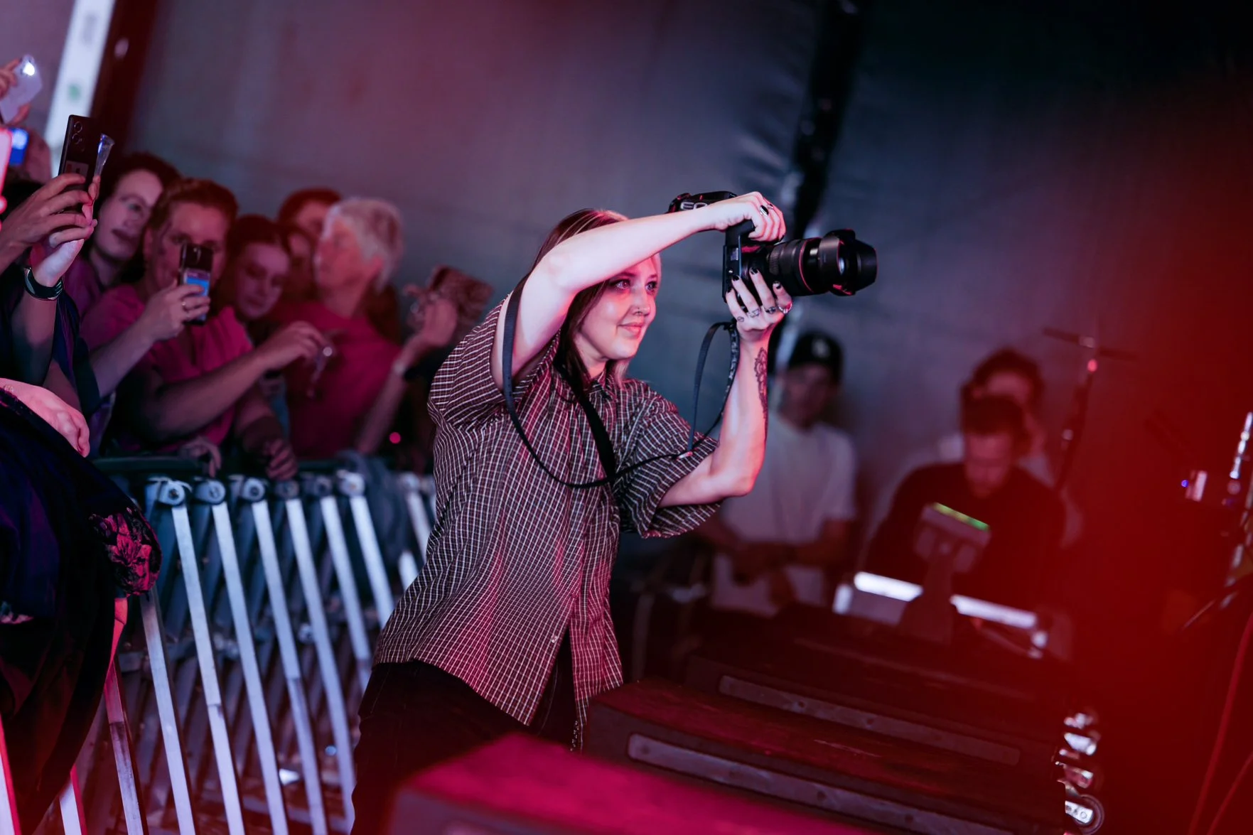 A woman with long hair taking a photo with a large camera at an indoor event, surrounded by a crowd of onlookers behind a metal barrier.