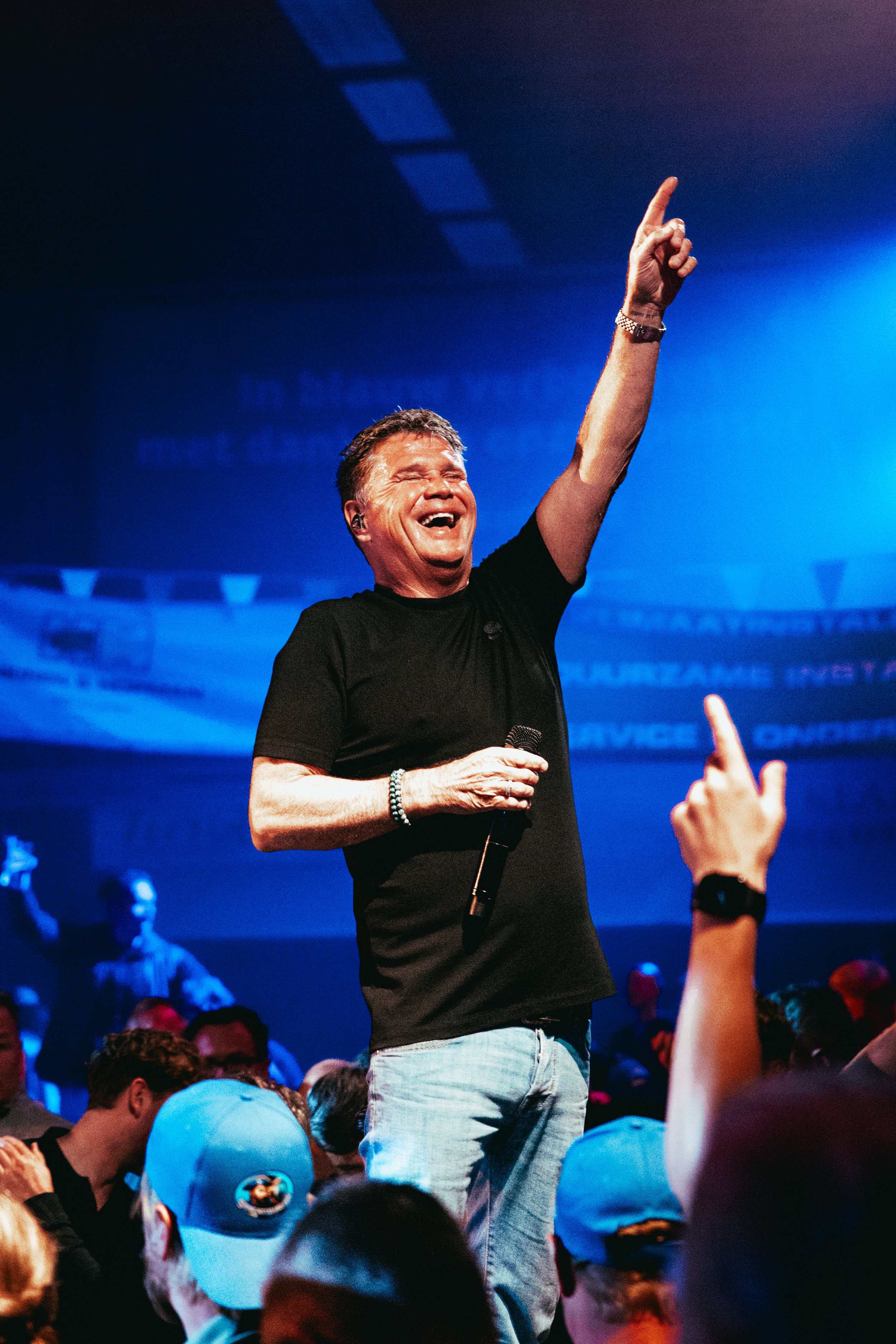 A man with short hair and light skin, smiling widely with eyes closed, is raising his right arm with his index finger pointing upward. He is holding a microphone in his left hand and wearing a black T-shirt, light jeans, a watch, and a beaded bracele