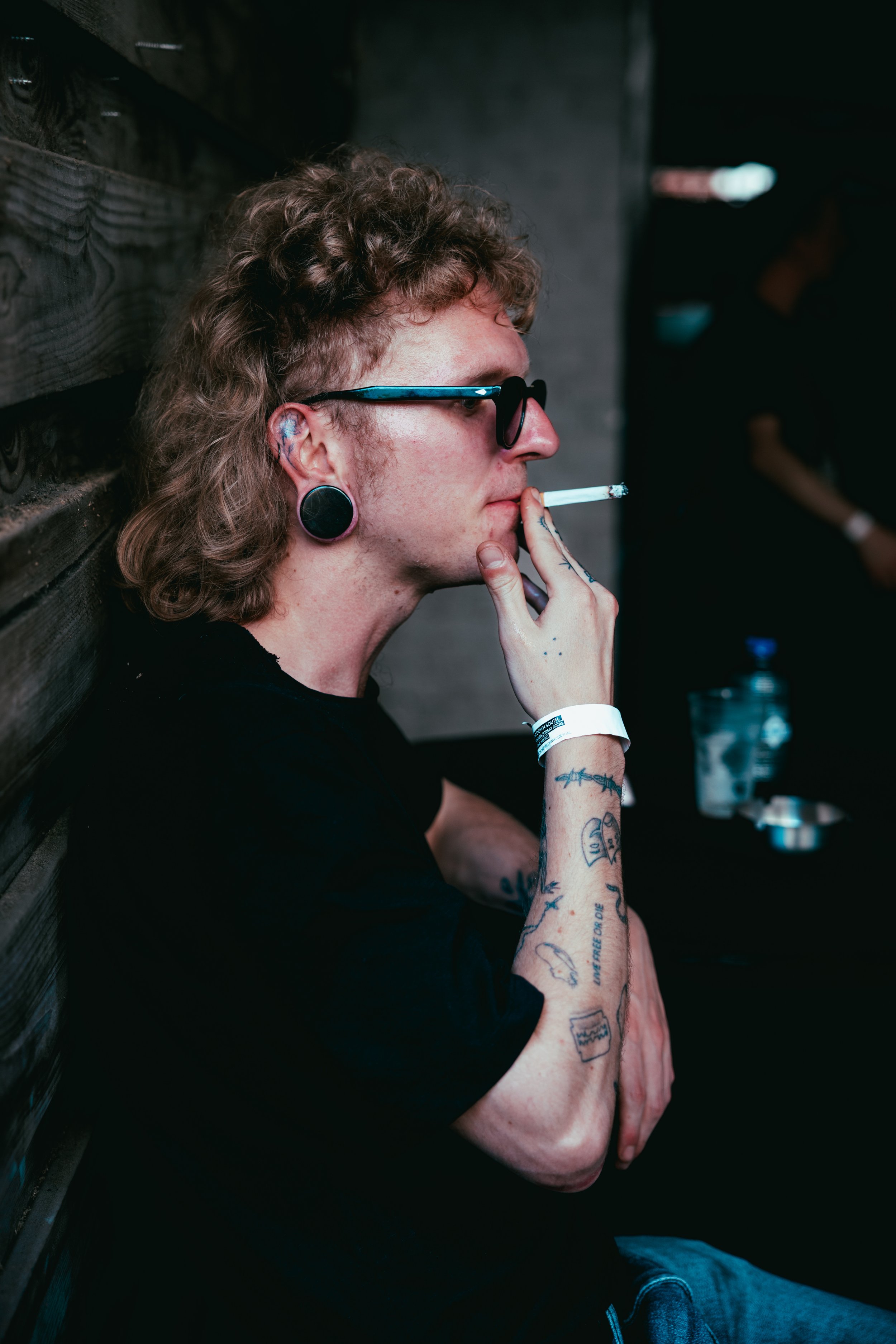 A person with curly hair, tattoos on their arm, large ear gauges, sunglasses, and a wristband, sitting with their back against a dark wall and smoking a cigarette.