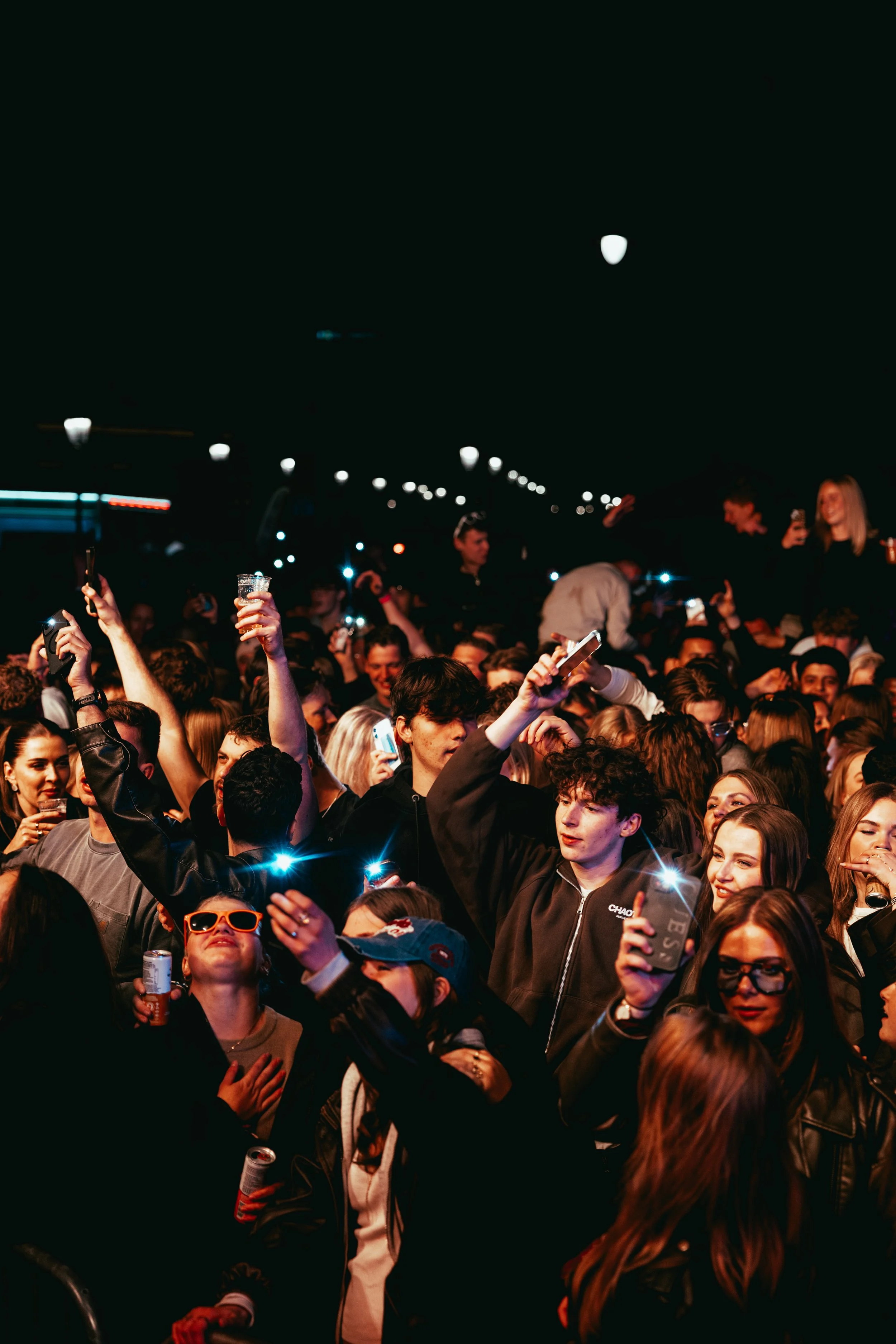 Crowd of people at night, many holding up phones and drinks, some smiling, in a lively social gathering.