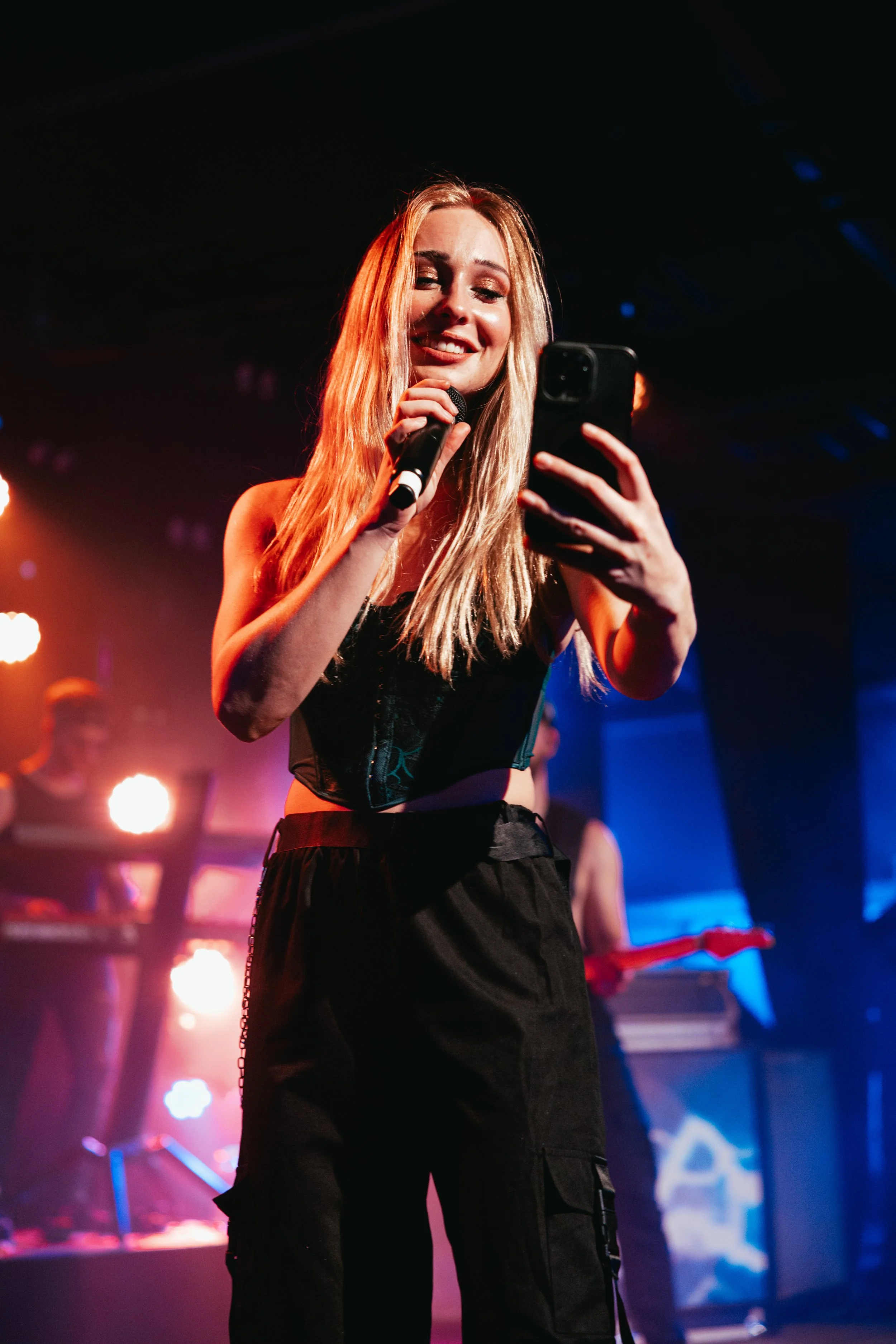 A young woman with long blonde hair is on stage with a microphone in one hand, looking at her phone with a smile. The background is dark with colorful stage lights and musical equipment.