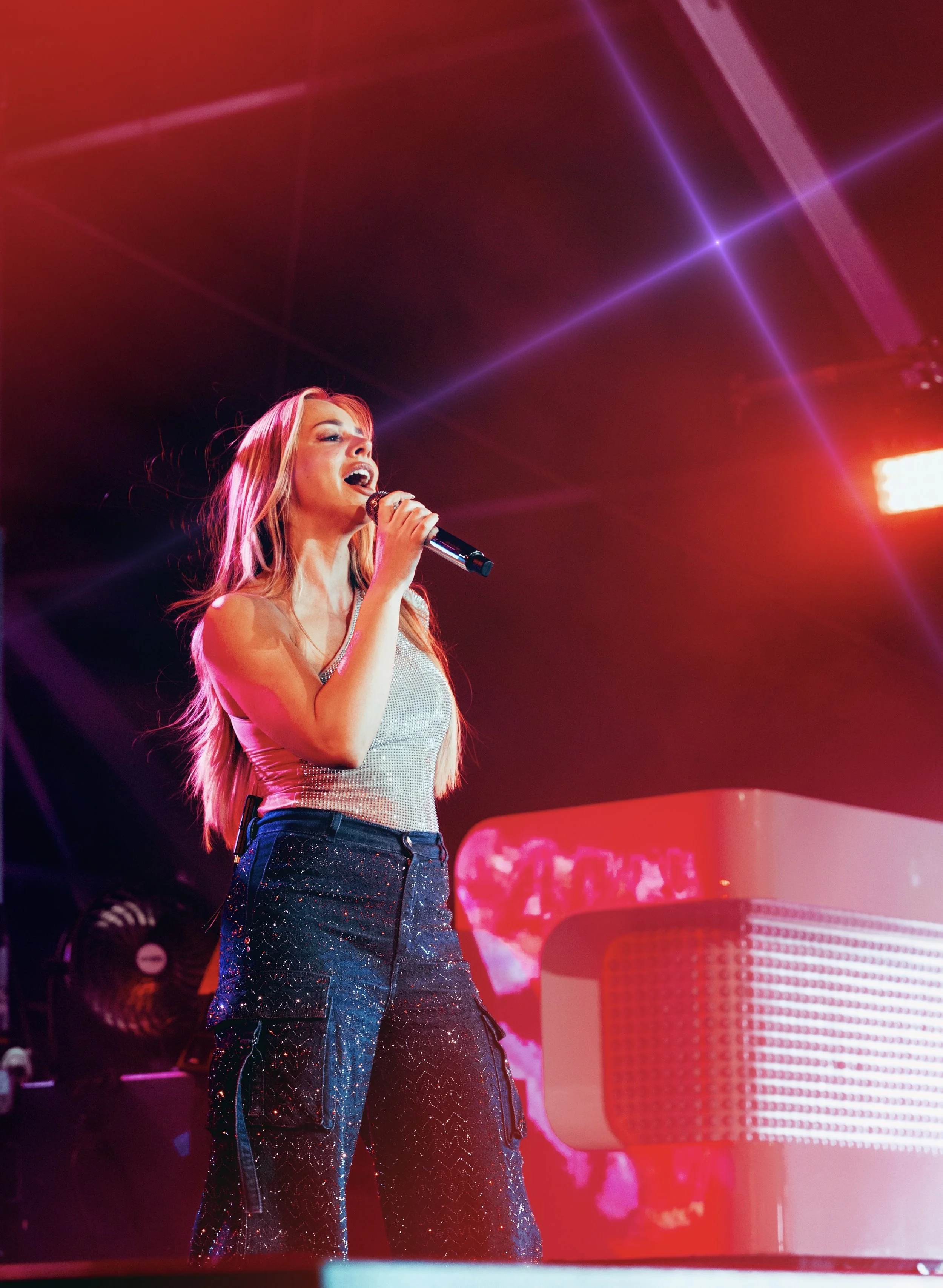 A young woman singing into a microphone on stage with colorful lighting and a red piano.
