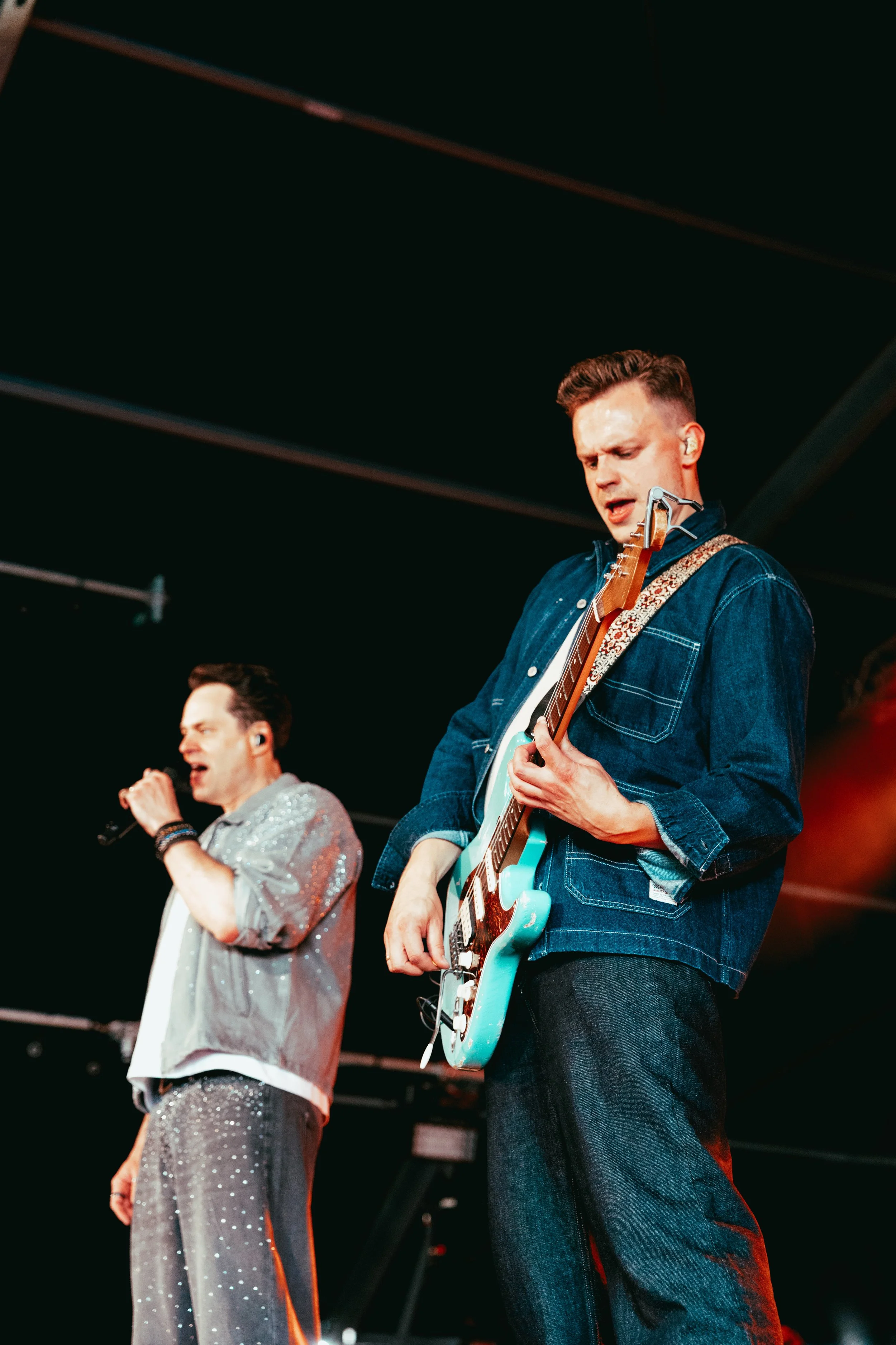 Two male musicians performing on stage. The man on the right is playing an electric guitar and wearing a denim jacket, while the man on the left is holding a microphone and wearing a shiny, silver, dotted outfit.