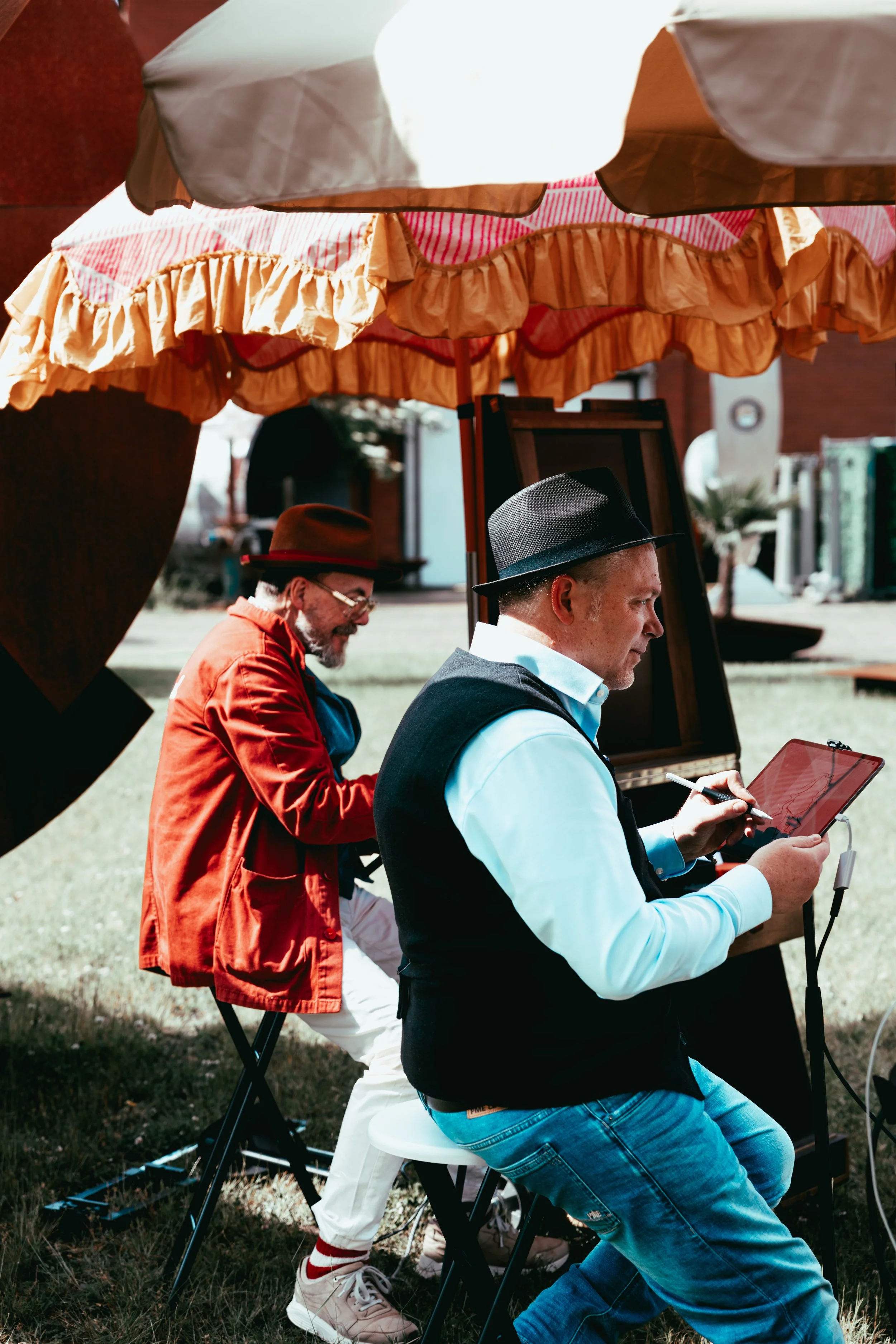 Two men sitting under a large patio umbrella, one wearing a black hat, light blue shirt, and vest, looking at a tablet; the other, wearing a brown hat and red jacket, looking down and laughing.