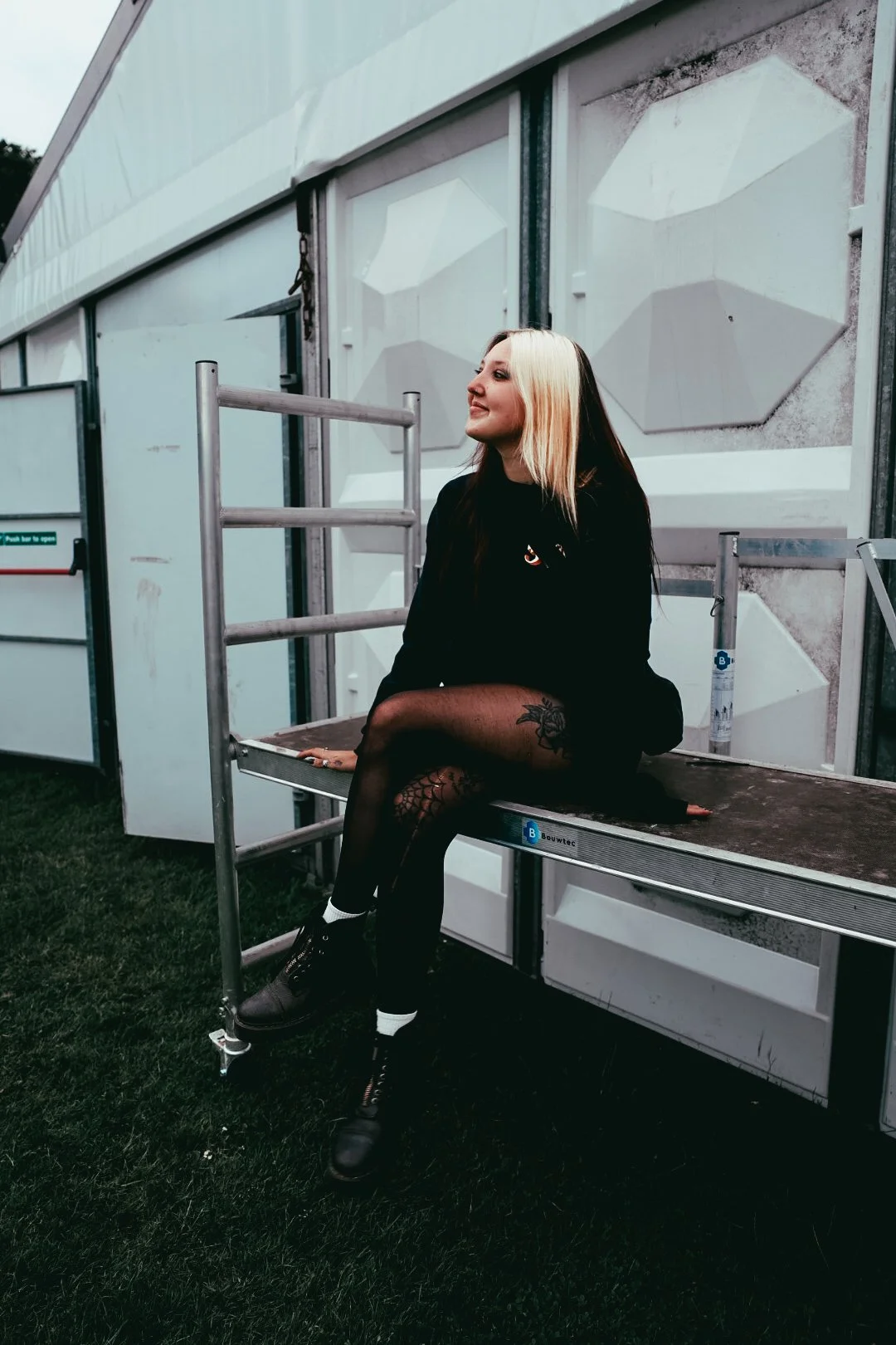 A woman with blonde and dark hair sitting on a platform outside a white building, dressed in black with fishnet stockings and black boots.