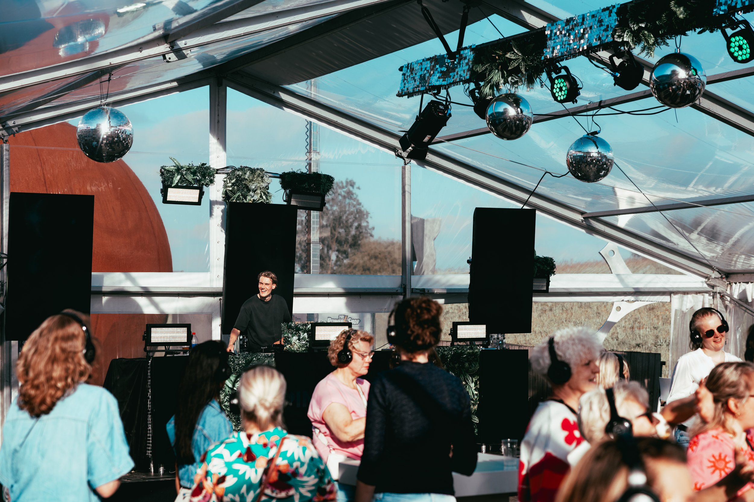 A DJ performing at an outdoor event with a crowd watching. The DJ is standing behind equipment, surrounded by people, with a tent covering and reflective disco balls hanging from the ceiling.