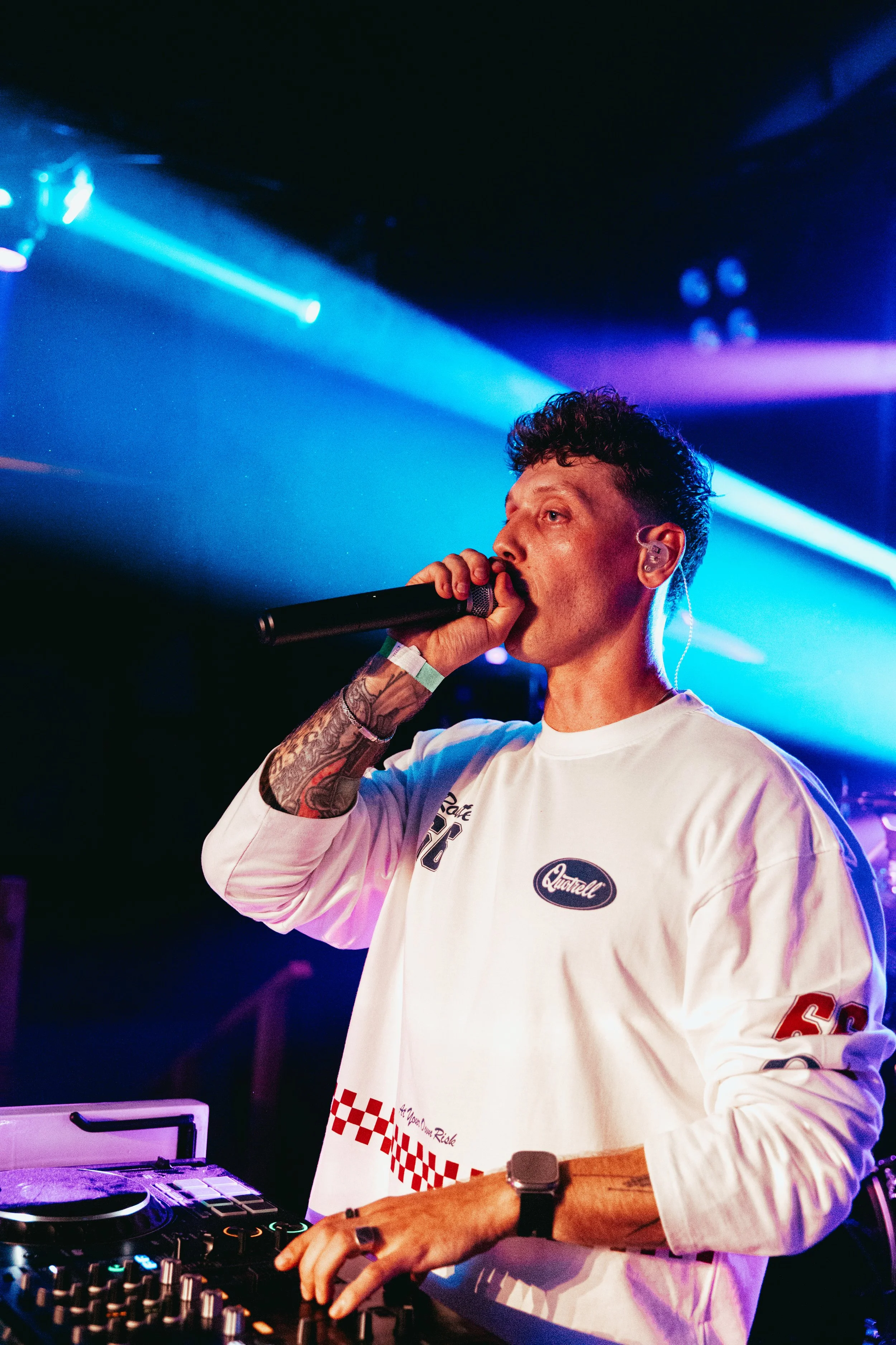 A DJ with short dark hair and tattoos on his arm, wearing a white shirt with red checking pattern and patches, is holding a microphone and standing in front of DJ equipment with colorful lighting around him.