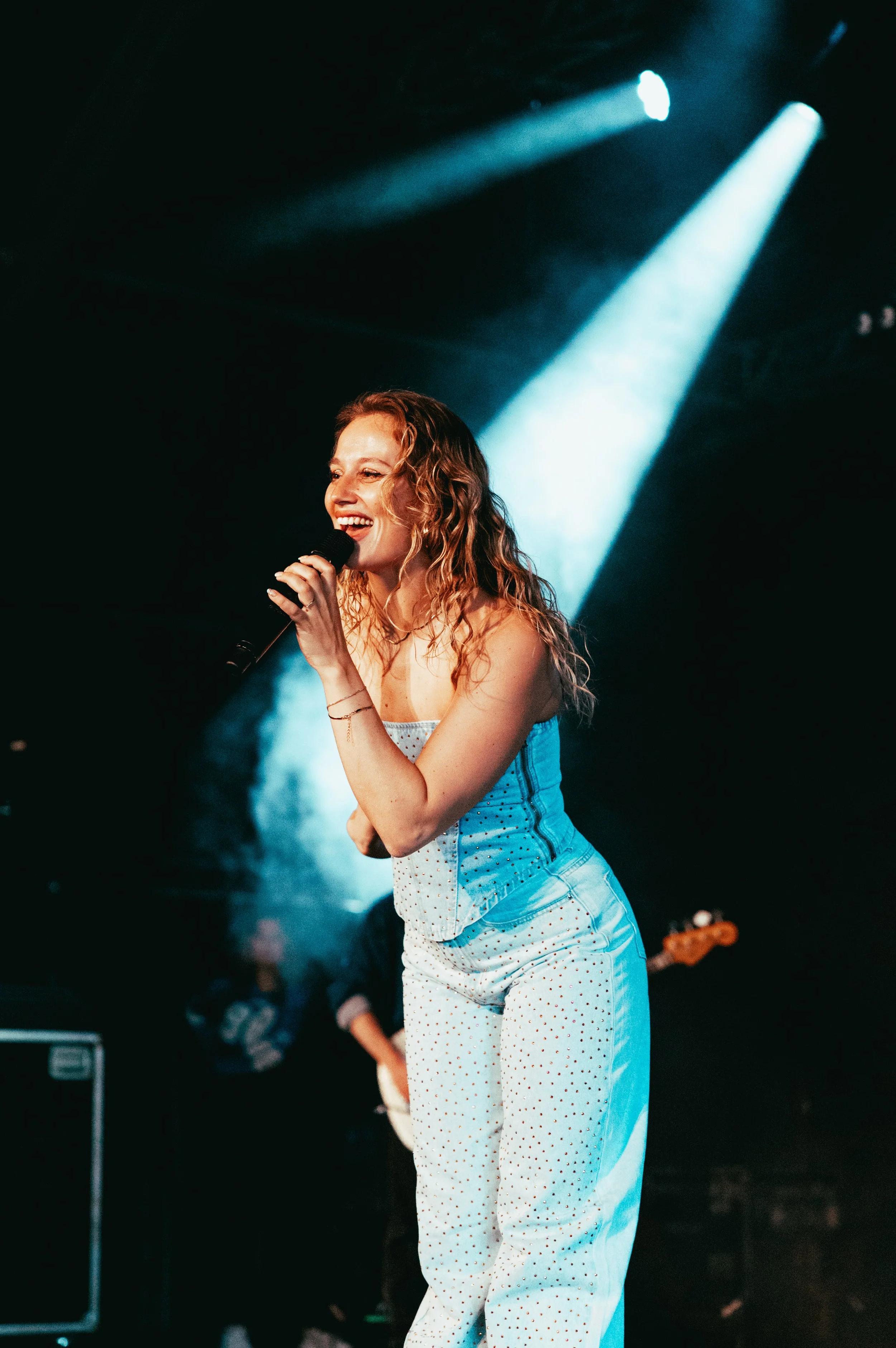 A woman with curly hair singing into a microphone on a stage with blue spotlights and a dark background.