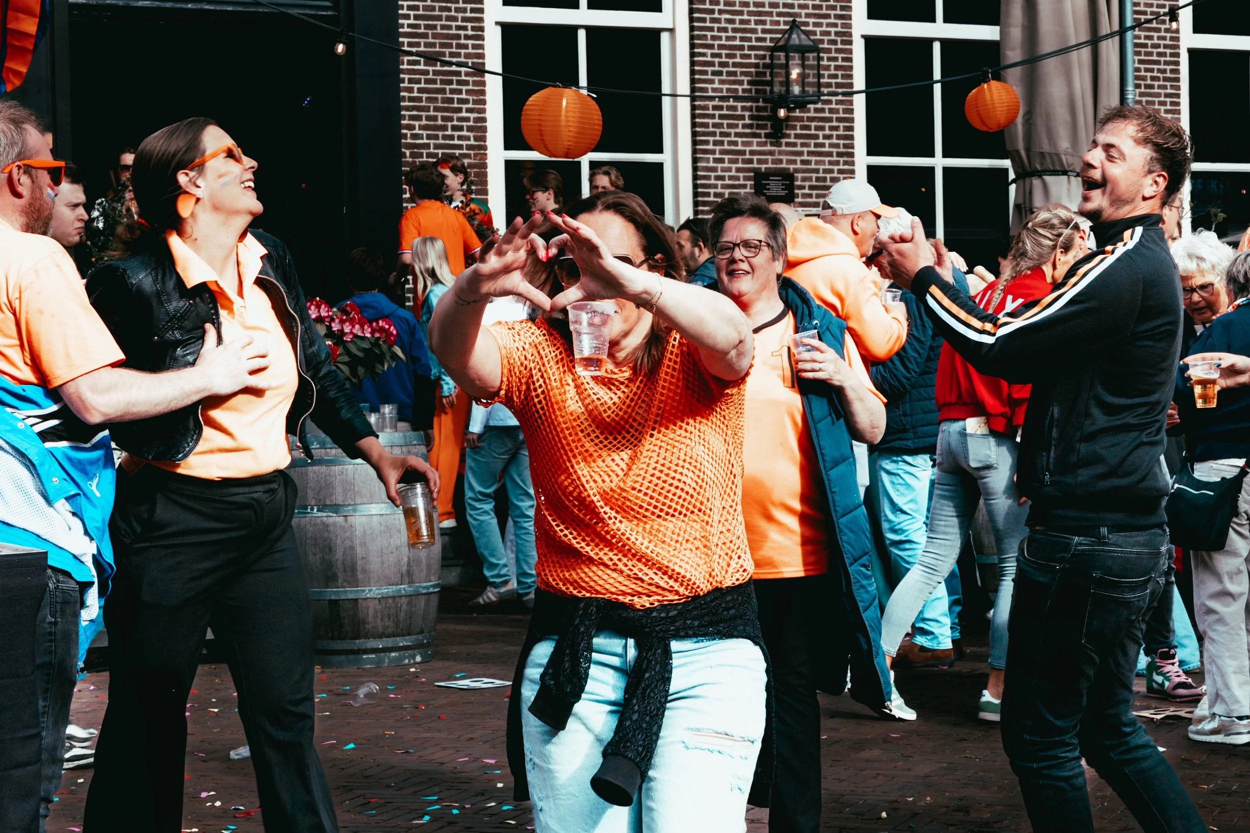 A group of people celebrating outdoors, wearing orange and black clothing, with paper lanterns hanging above them, and a brick building in the background.